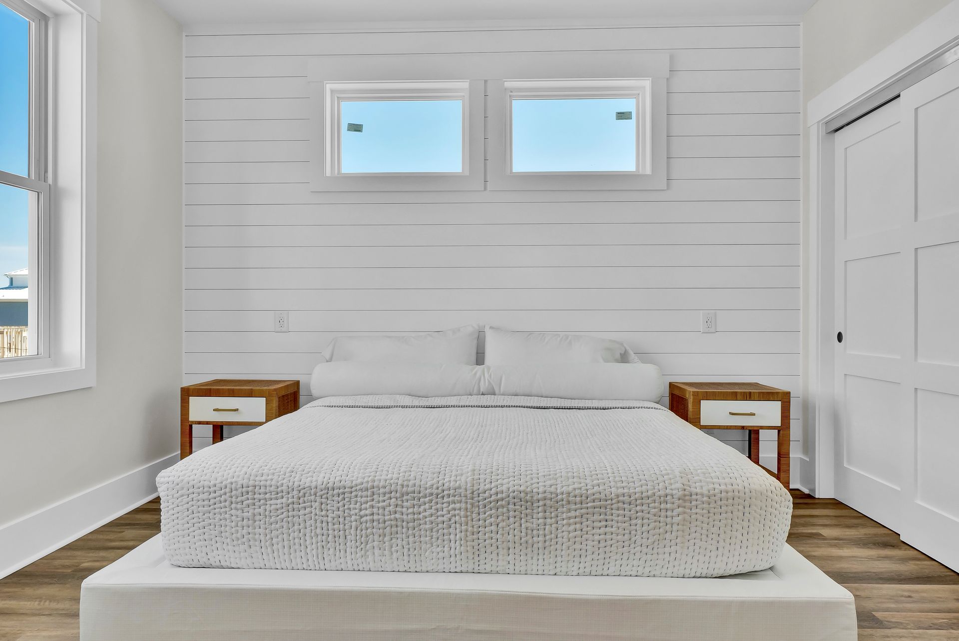 Bedroom with a white bed, shiplap wall, wooden nightstands, and two small windows.