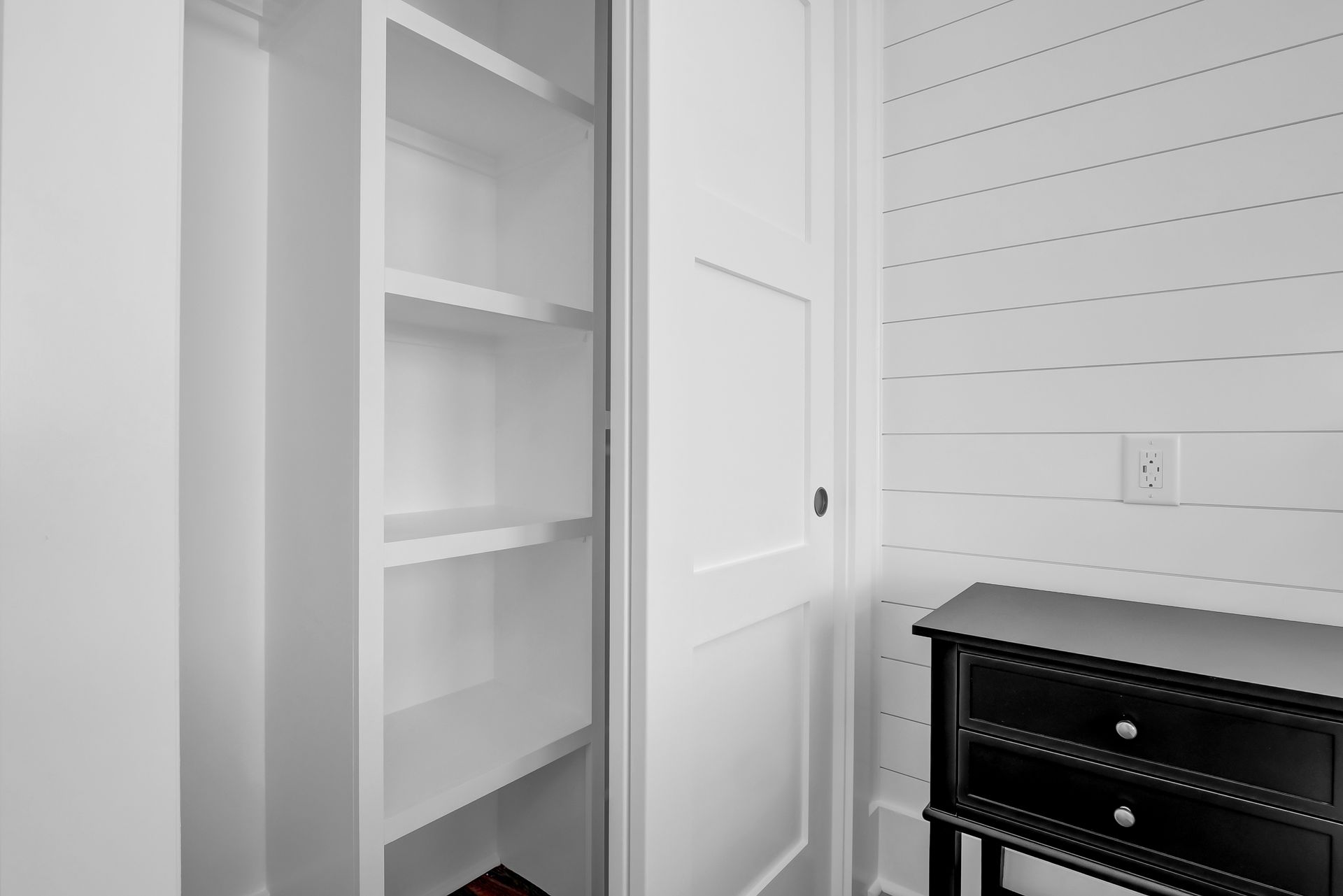 White built-in shelves next to a white door, next to a black nightstand with two drawers and an outlet.