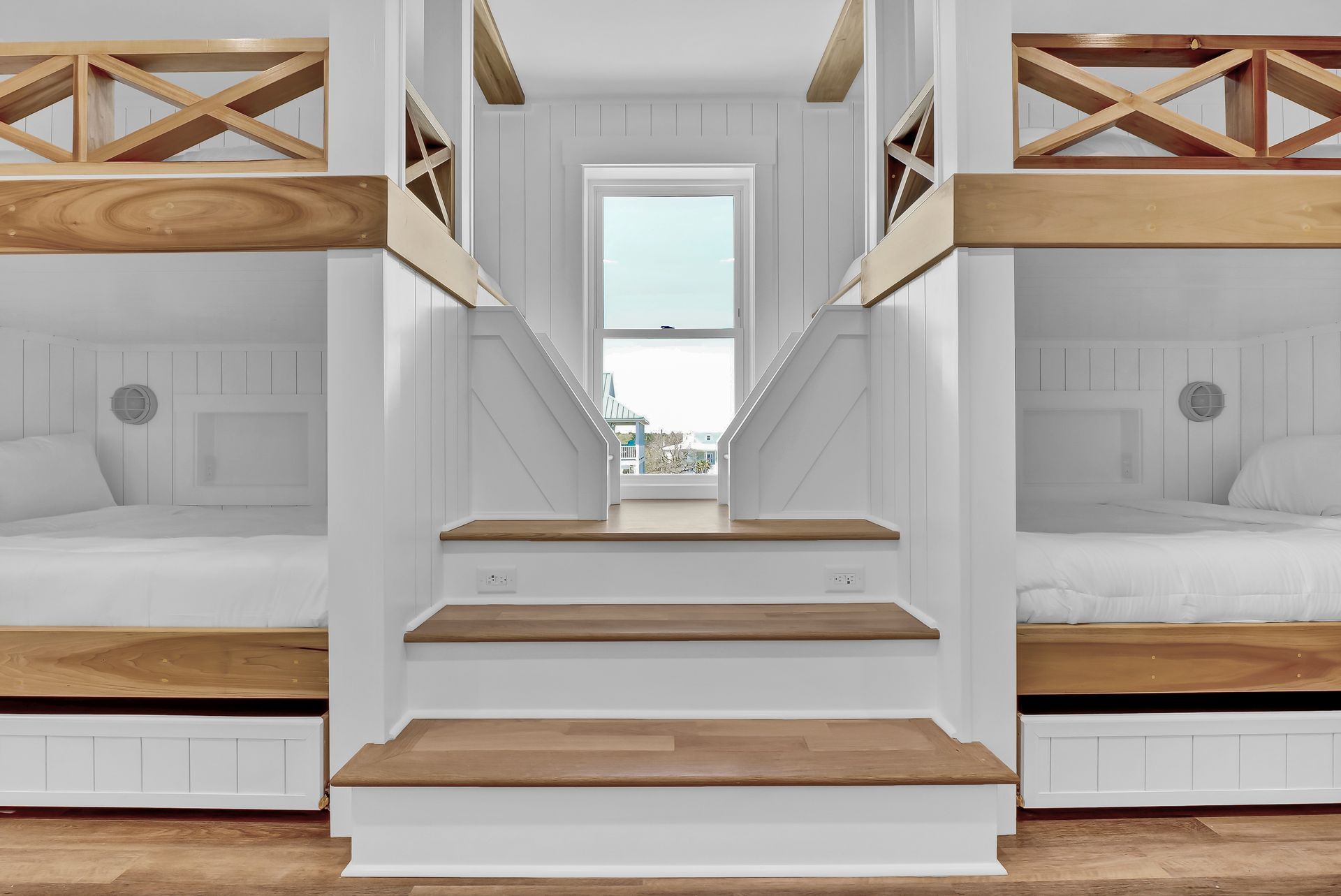 Bunk bed room with wood accents. Two beds on either side of stairs leading to a window. White walls.