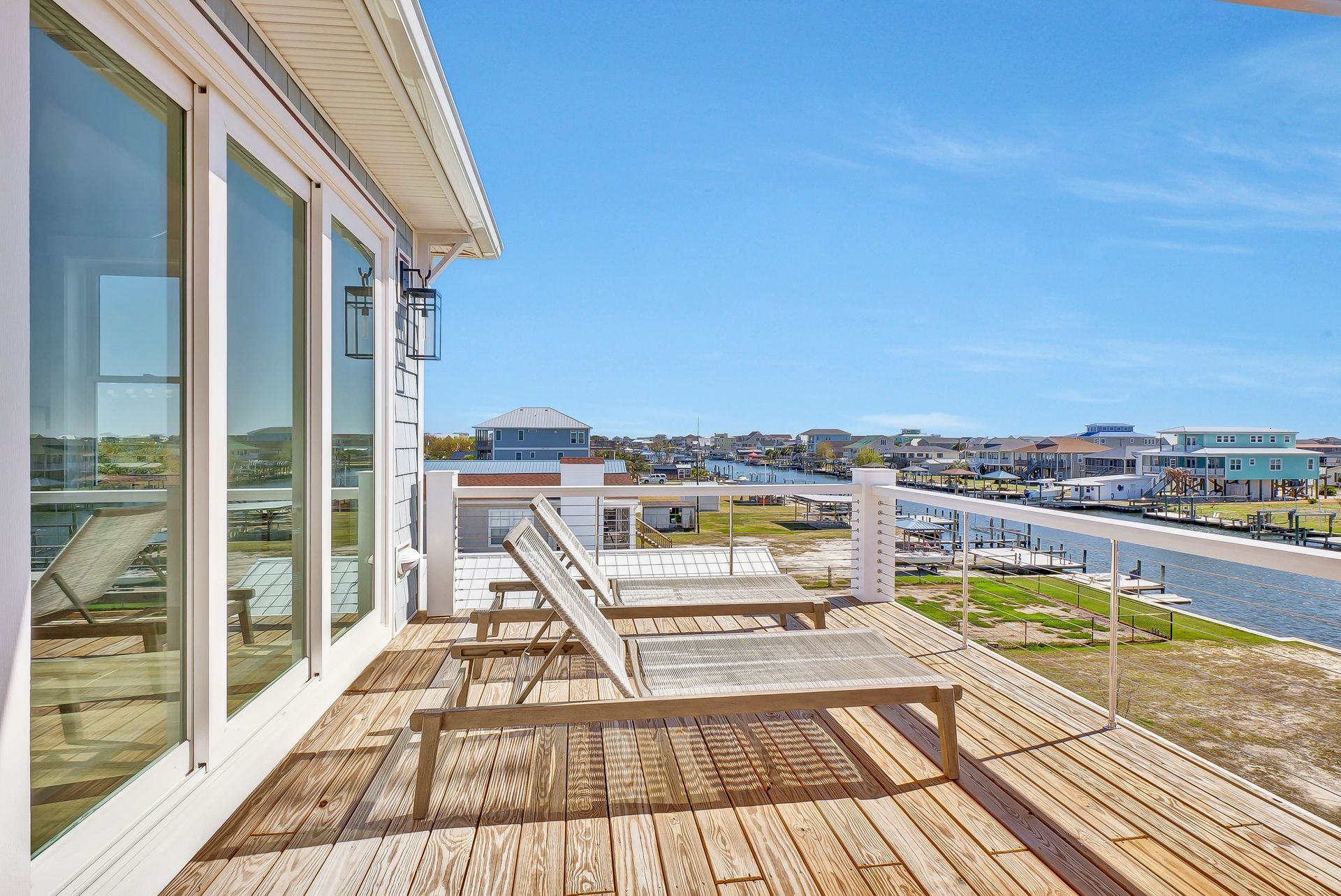 Wooden deck with two lounge chairs, overlooking a canal and houses under a bright blue sky.