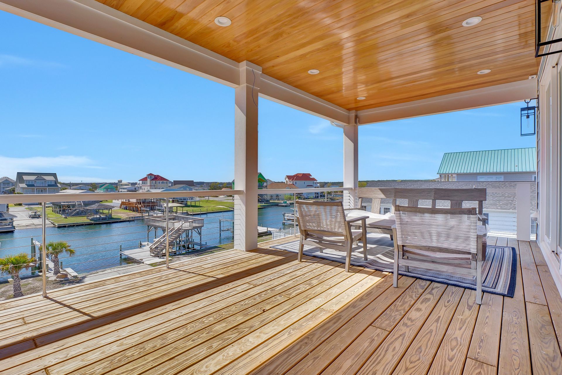 Wooden deck overlooking water, furnished with chairs, under a wood ceiling. Sunny day, blue sky.