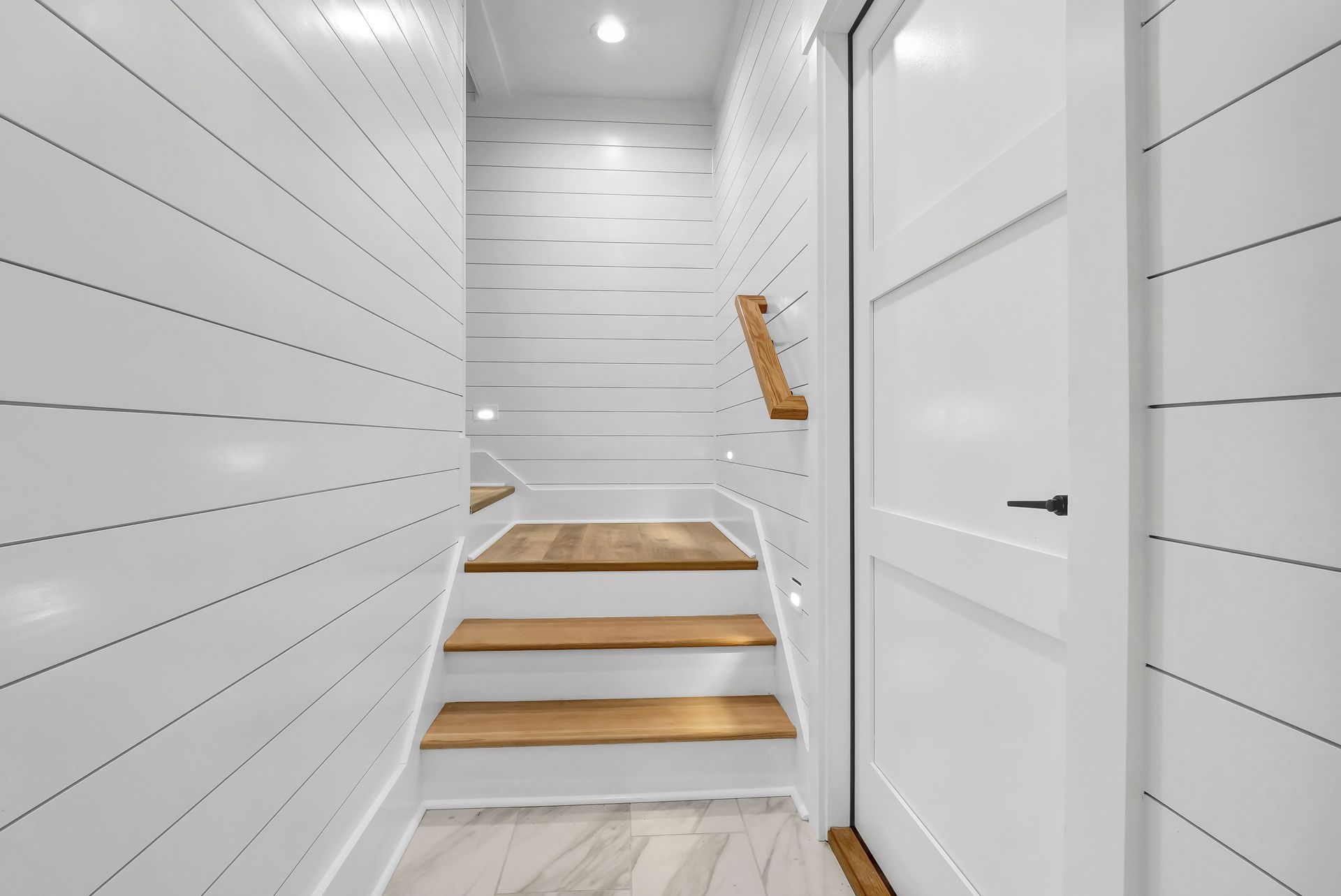 White shiplap hallway with wooden stairs and door. Overhead lights illuminate the space.