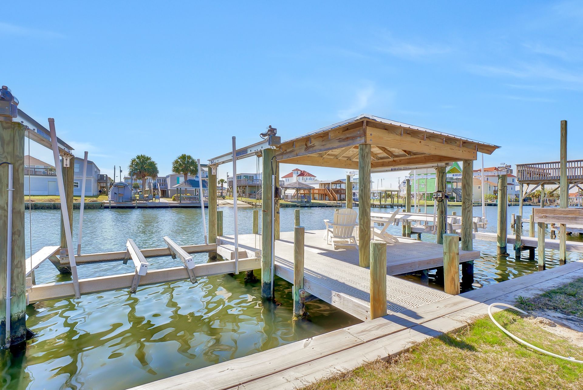 Wooden dock with a canopy over a seating area, canal with houses in the background, blue sky.