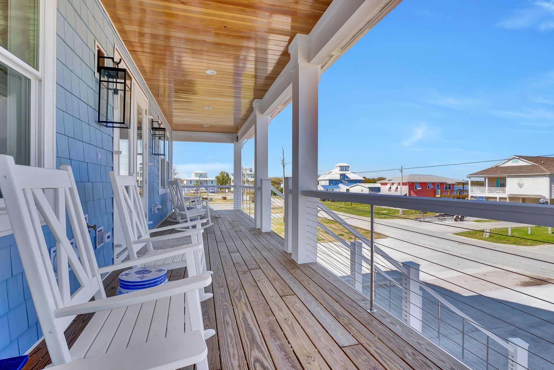 Covered porch with white rocking chairs, blue siding, wood ceiling and deck, overlooking a street and other buildings.