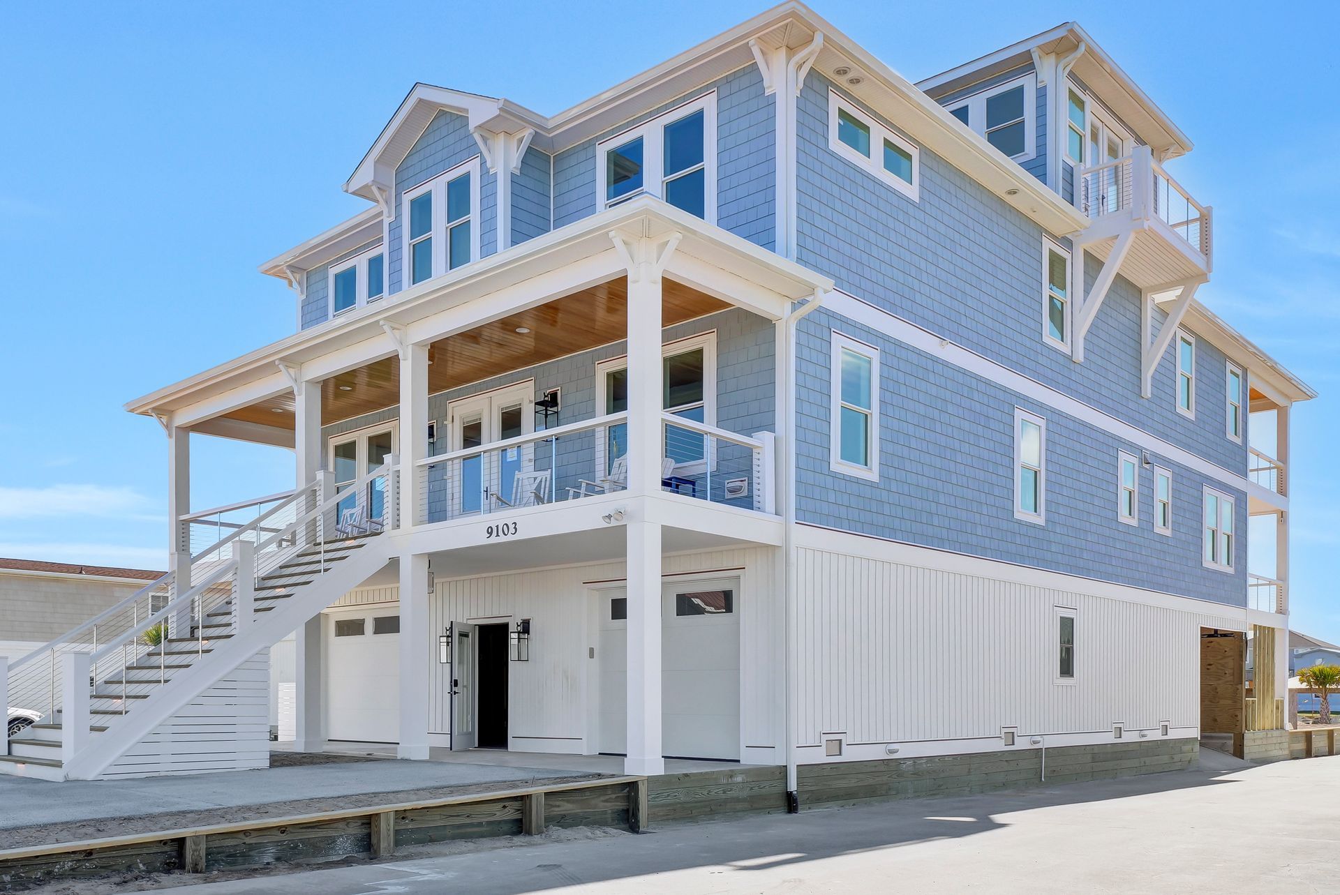 Blue and white multi-story house with decks, stairs, and garage doors against a clear blue sky.