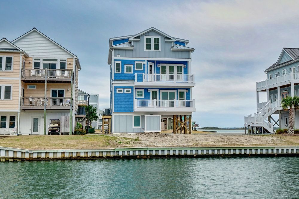 Three-story blue and grey coastal home on a waterway, with balconies, and surrounded by similar homes.