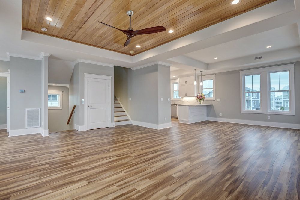 Spacious living area with wood flooring and ceiling, white walls, and a view of the kitchen and windows.