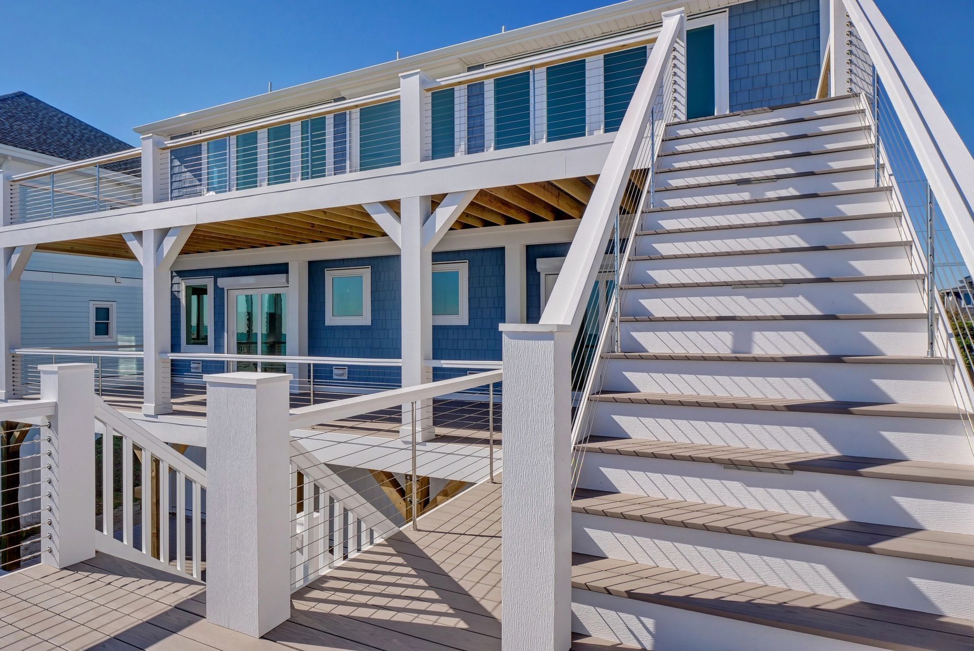 White stairway and deck leading to a blue beach house with multiple windows and a sunny blue sky.
