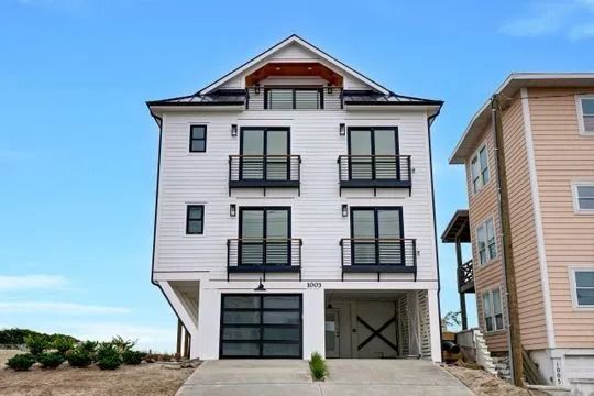 White multi-story house with black trim, balconies, and garage doors, blue sky background.