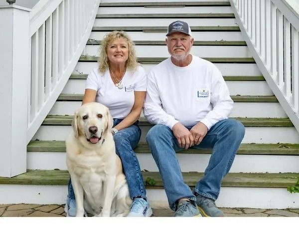 Couple and dog sitting on outdoor steps. Man in cap, woman smiling, yellow lab beside them.