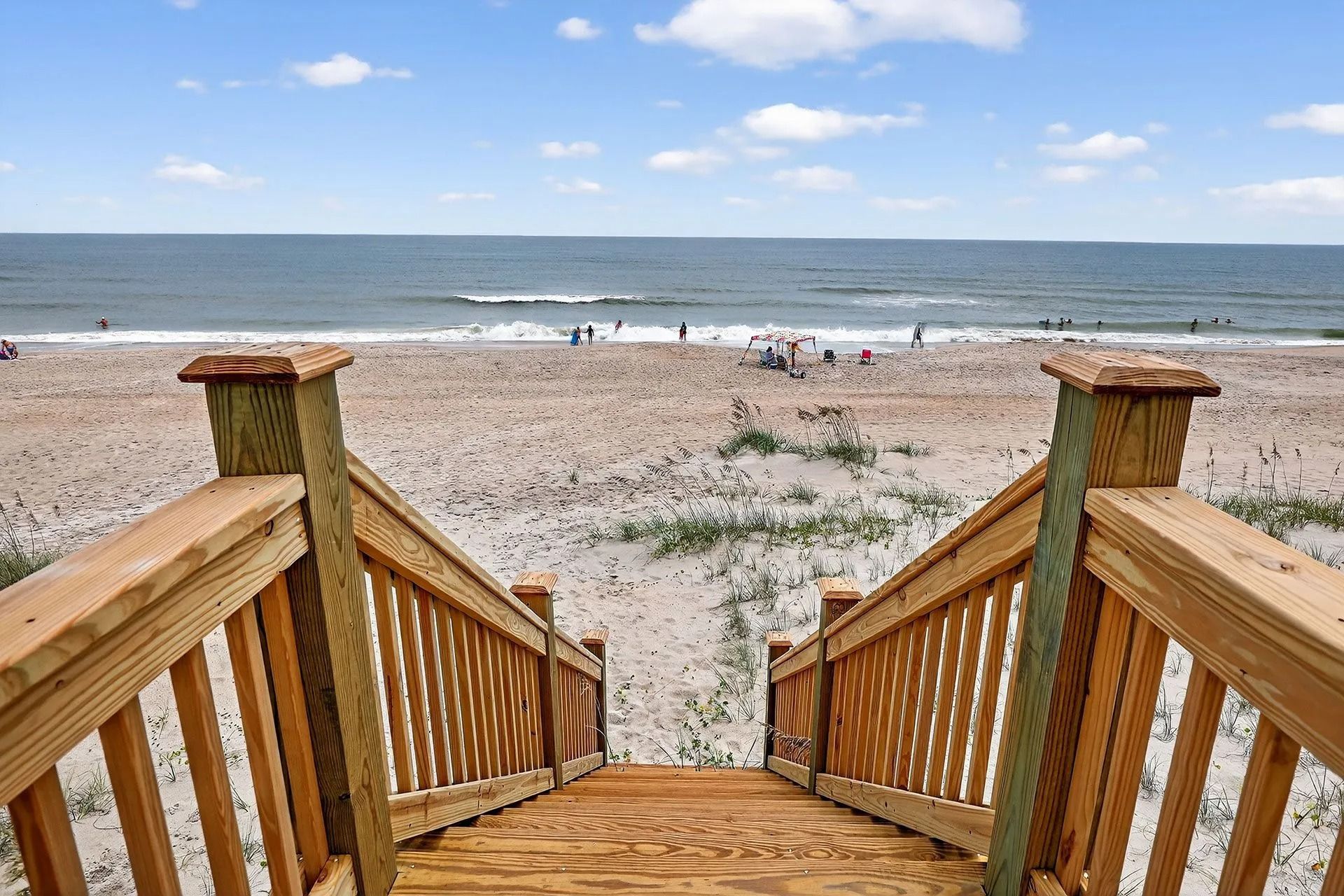 Wooden stairs leading to a sandy beach and ocean under a blue sky. People are visible on the beach.