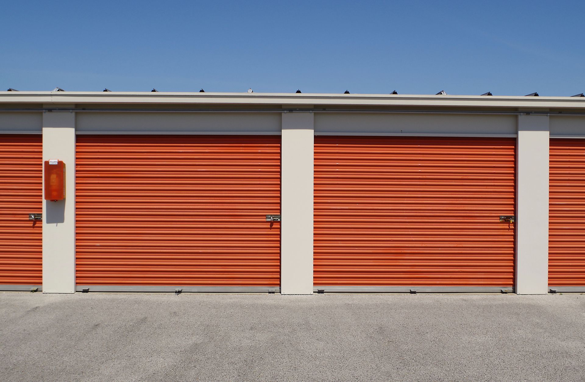Orange storage unit doors between white pillars on a gravel lot under a clear blue sky.