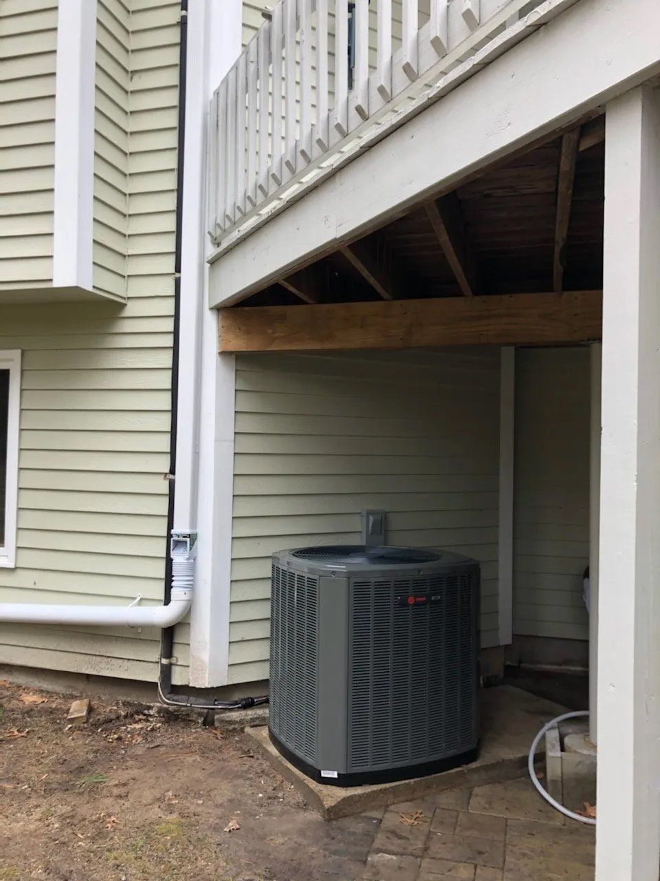 An air conditioner is sitting under a deck next to a house.