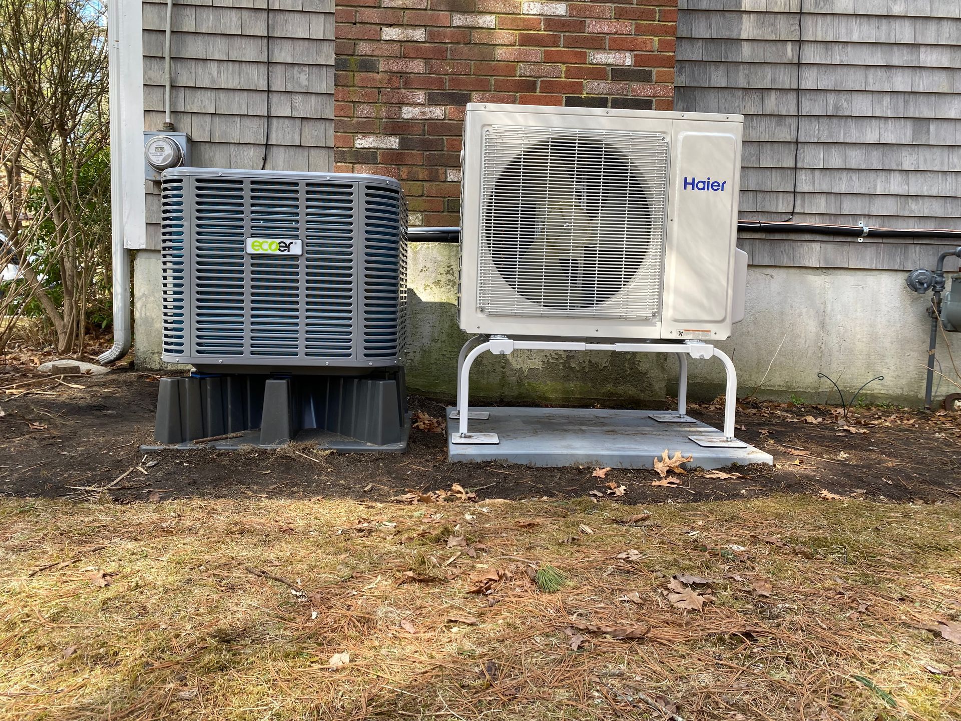 Two air conditioners are sitting next to each other on the side of a house.