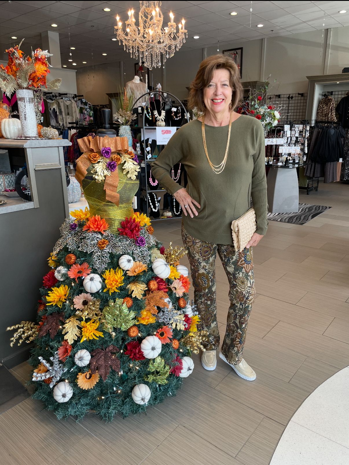 Woman poses by a large floral display, possibly inside a shop. She wears green and camo.