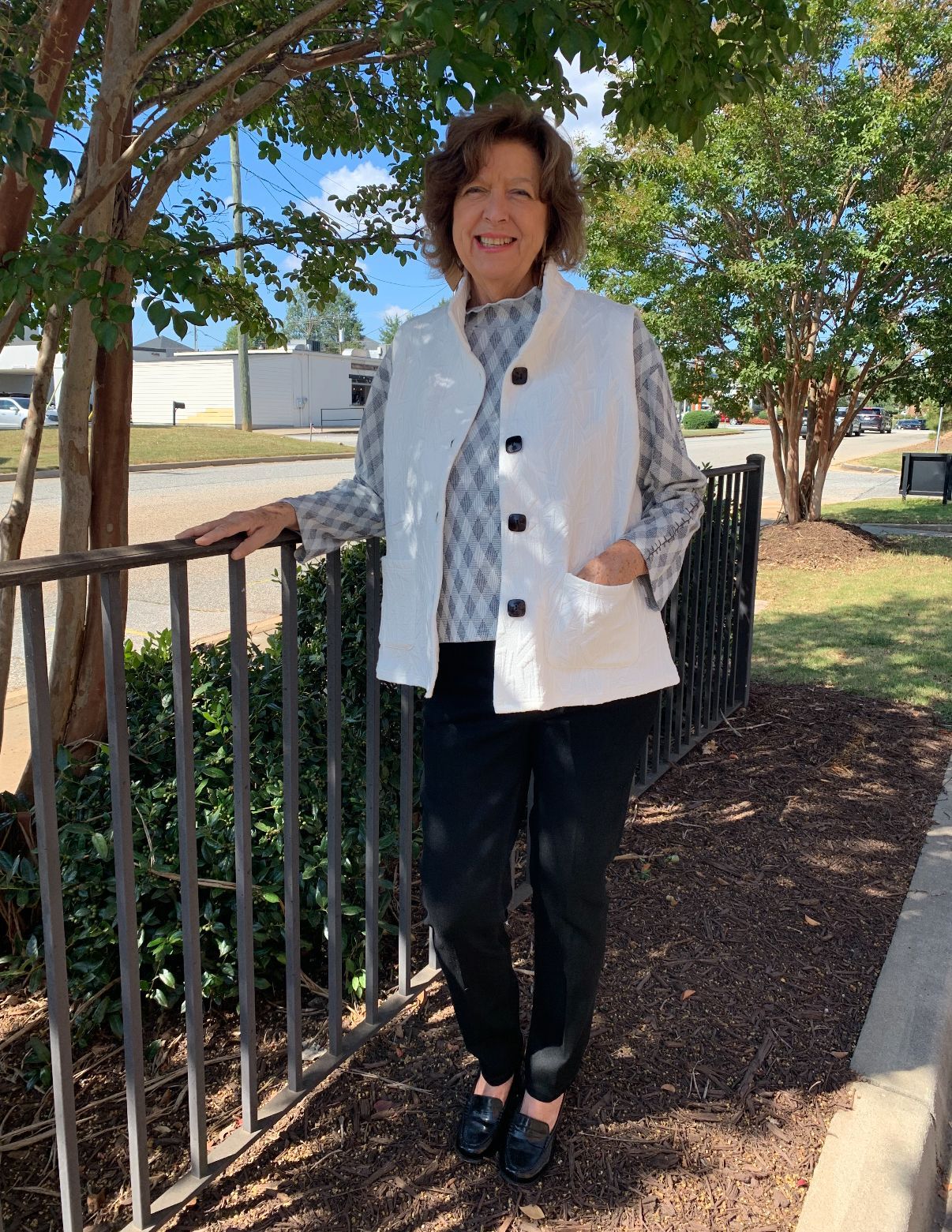 Woman in white vest, patterned shirt, black pants, and loafers smiles outdoors near a black fence.