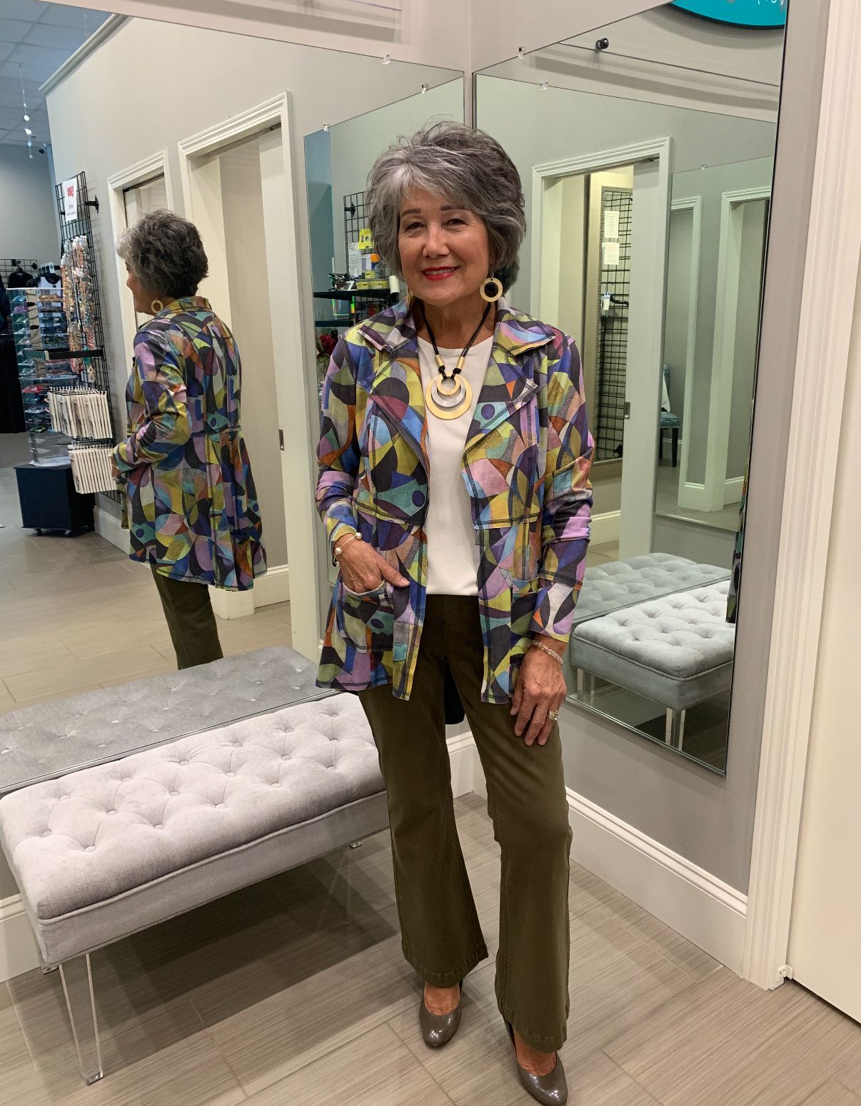 Woman in a colorful jacket and olive pants, smiling in a store, standing by a mirror.
