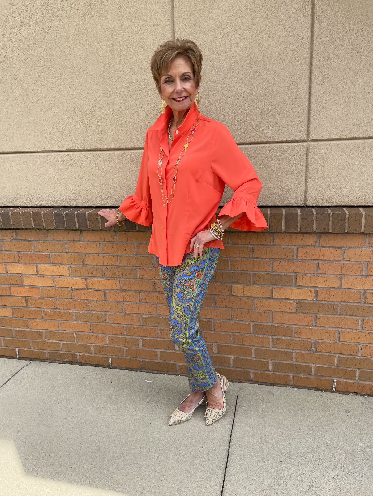 Woman wearing orange shirt, floral pants, standing outdoors.