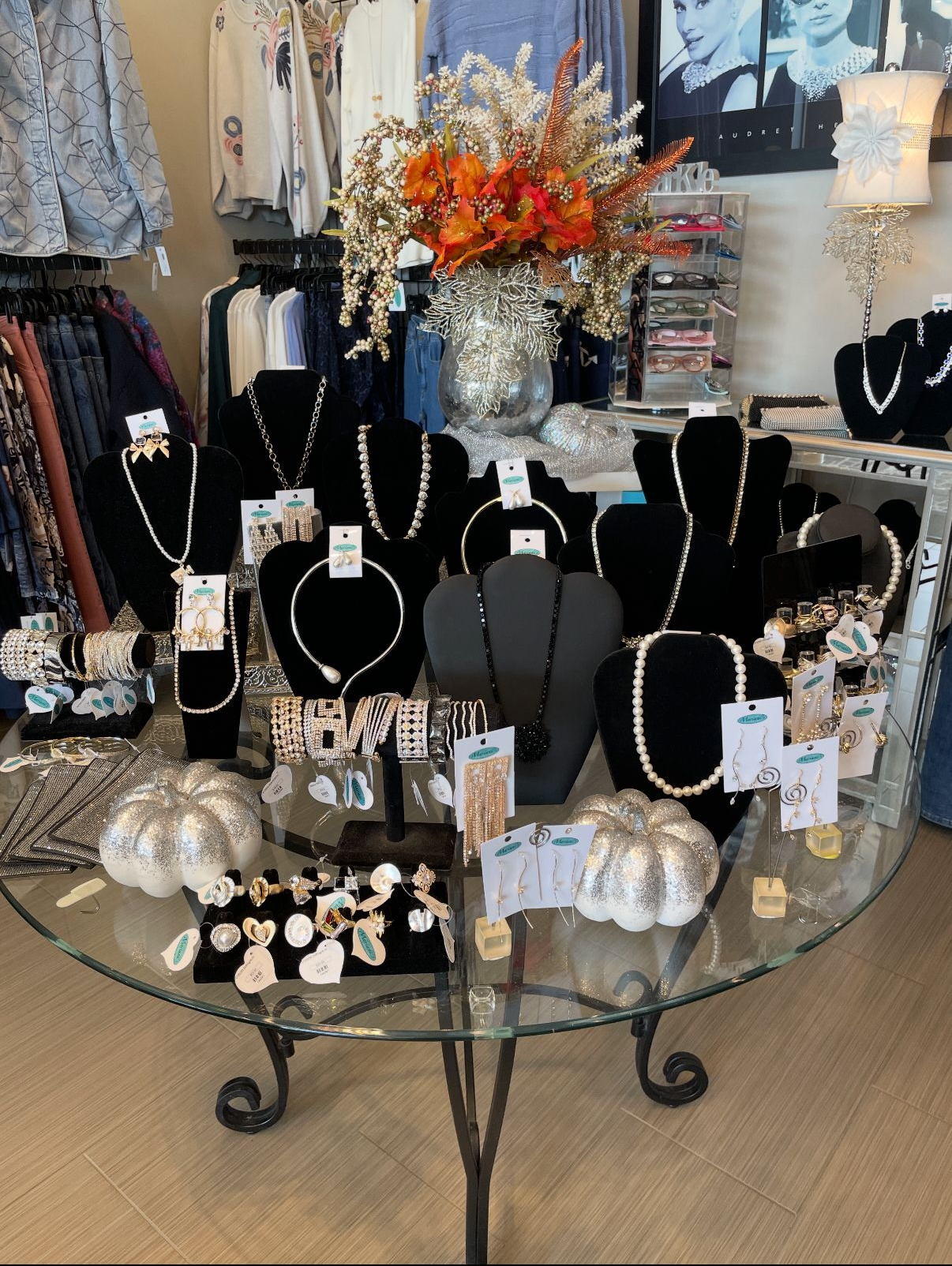 Jewelry display on a glass table with necklaces and earrings. Flowers and shop interior in background.