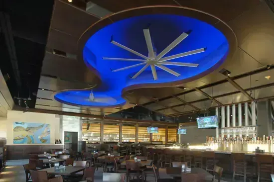 Restaurant interior with blue-lit ceiling design and large fan. Tables, bar, and open windows are visible.