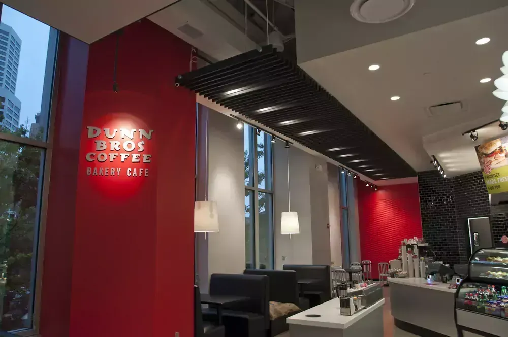 A modern restaurant storefront with glass walls; green logo, seating visible inside, and wooden light fixture.