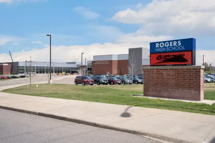 Rogers High School building with sign, cars in parking lot, and grassy area under a blue sky.