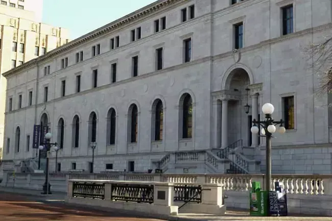 Large, light-colored building with arched windows, steps, and a classical facade. Street-level view, sunny day.
