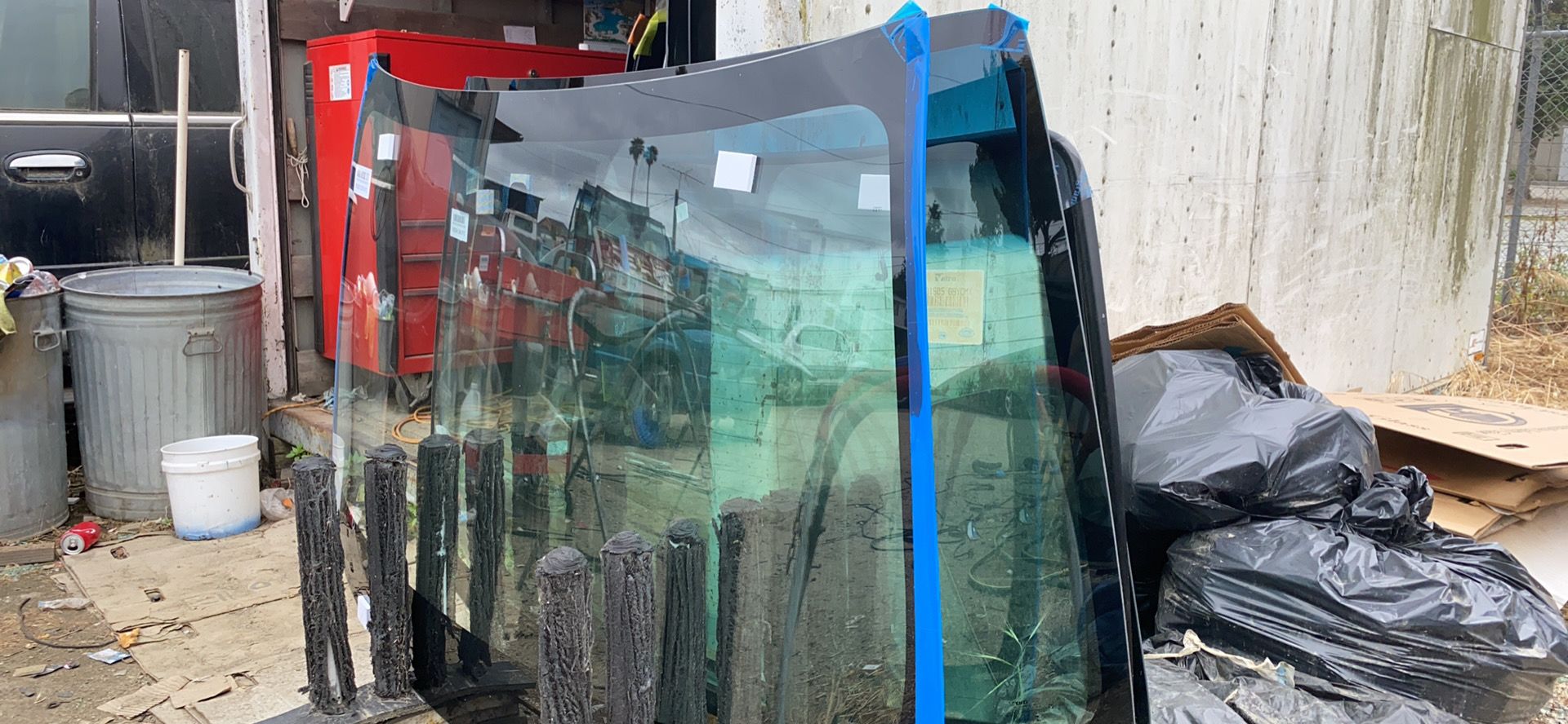 Car windshields stacked against a concrete wall, held together with blue tape. Trash bags and a garbage can are nearby.