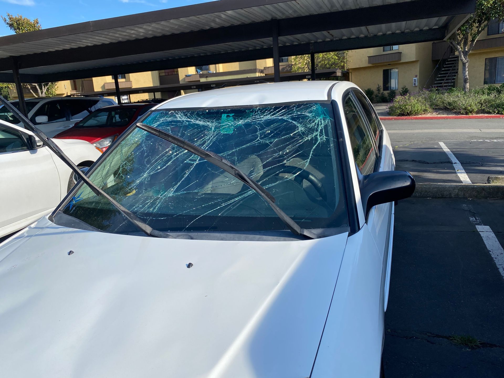 White car with a severely cracked windshield parked under a covered parking area.