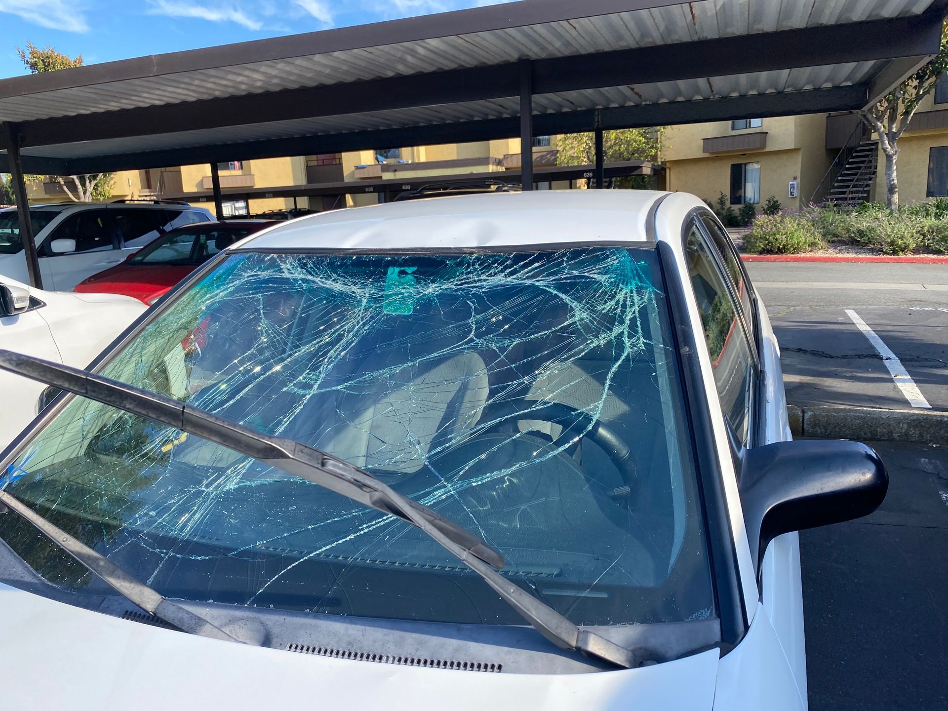 White car with severely cracked windshield parked under a carport.