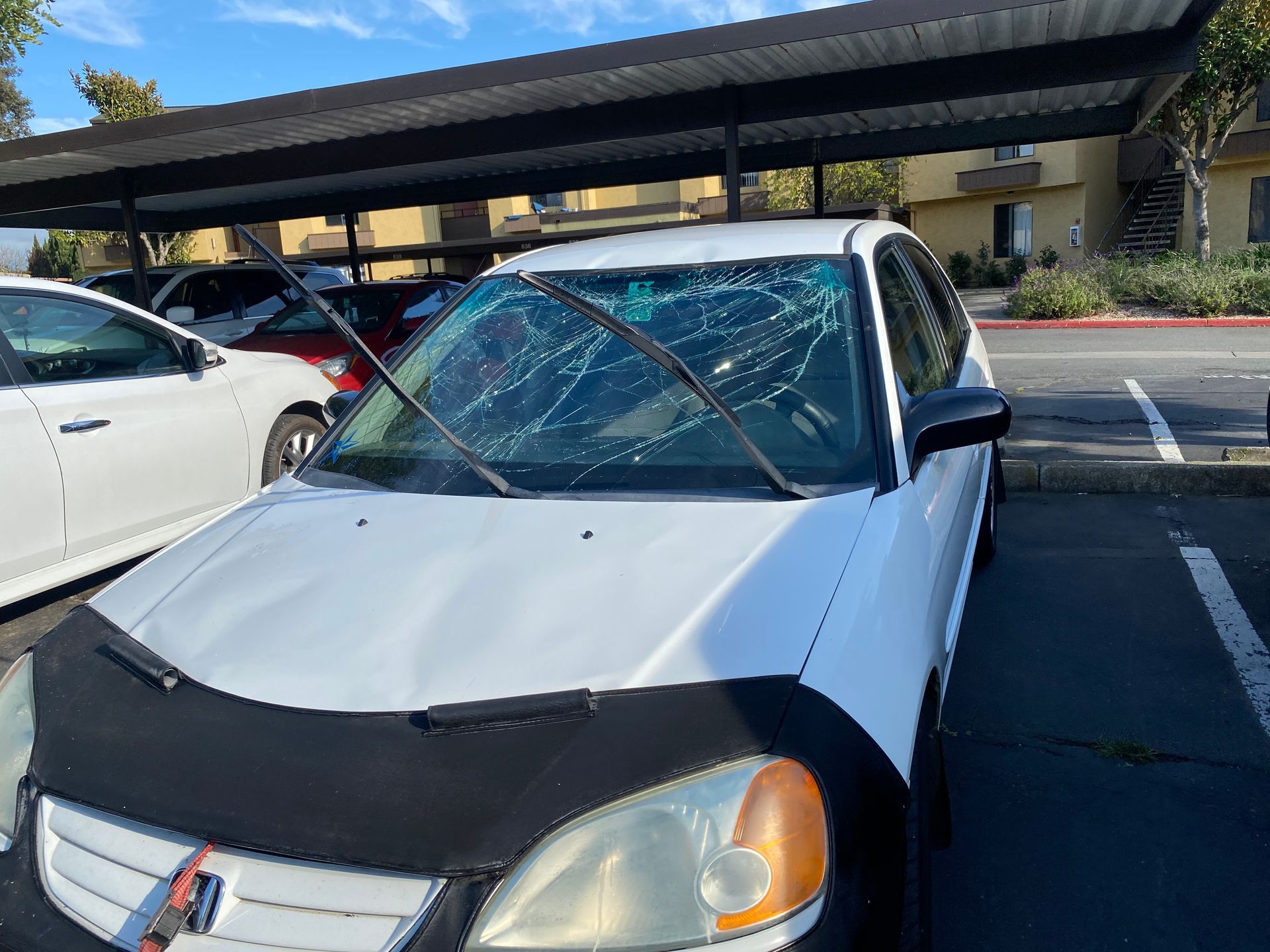 White car with shattered windshield parked under a carport.