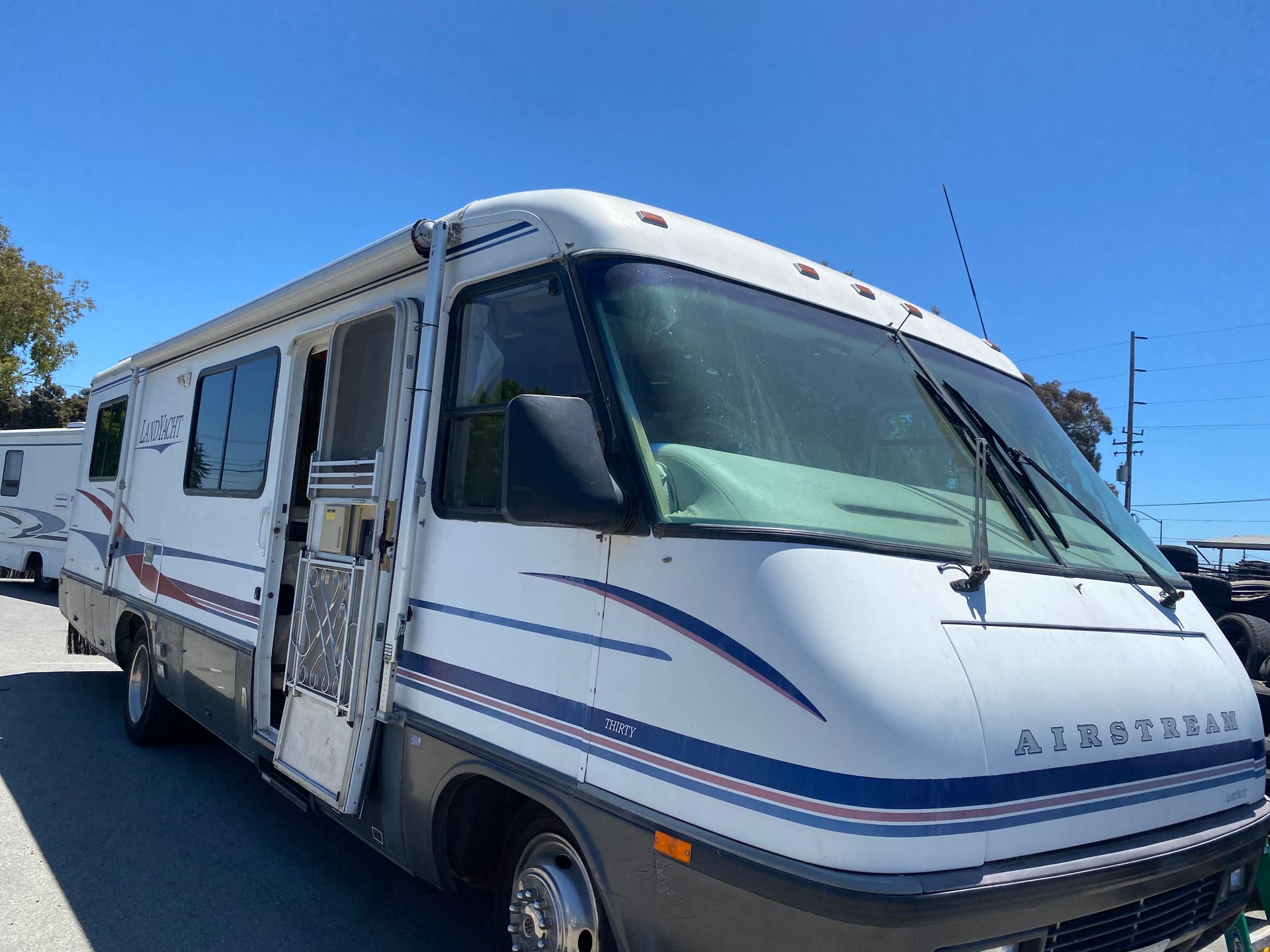 White RV with blue and red stripes, parked outside under a blue sky, door open.