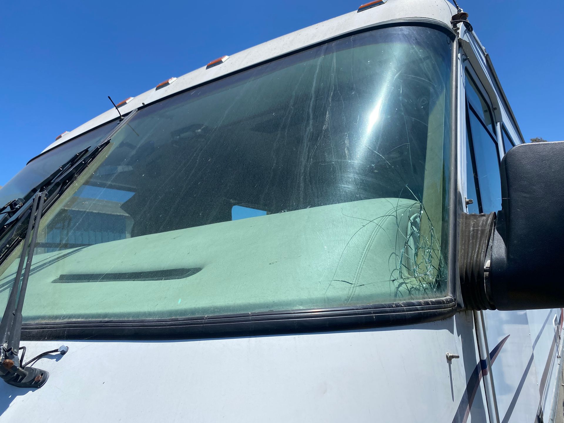 Close-up of the front of a white RV with a dirty windshield and driver's side mirror, under a clear blue sky.