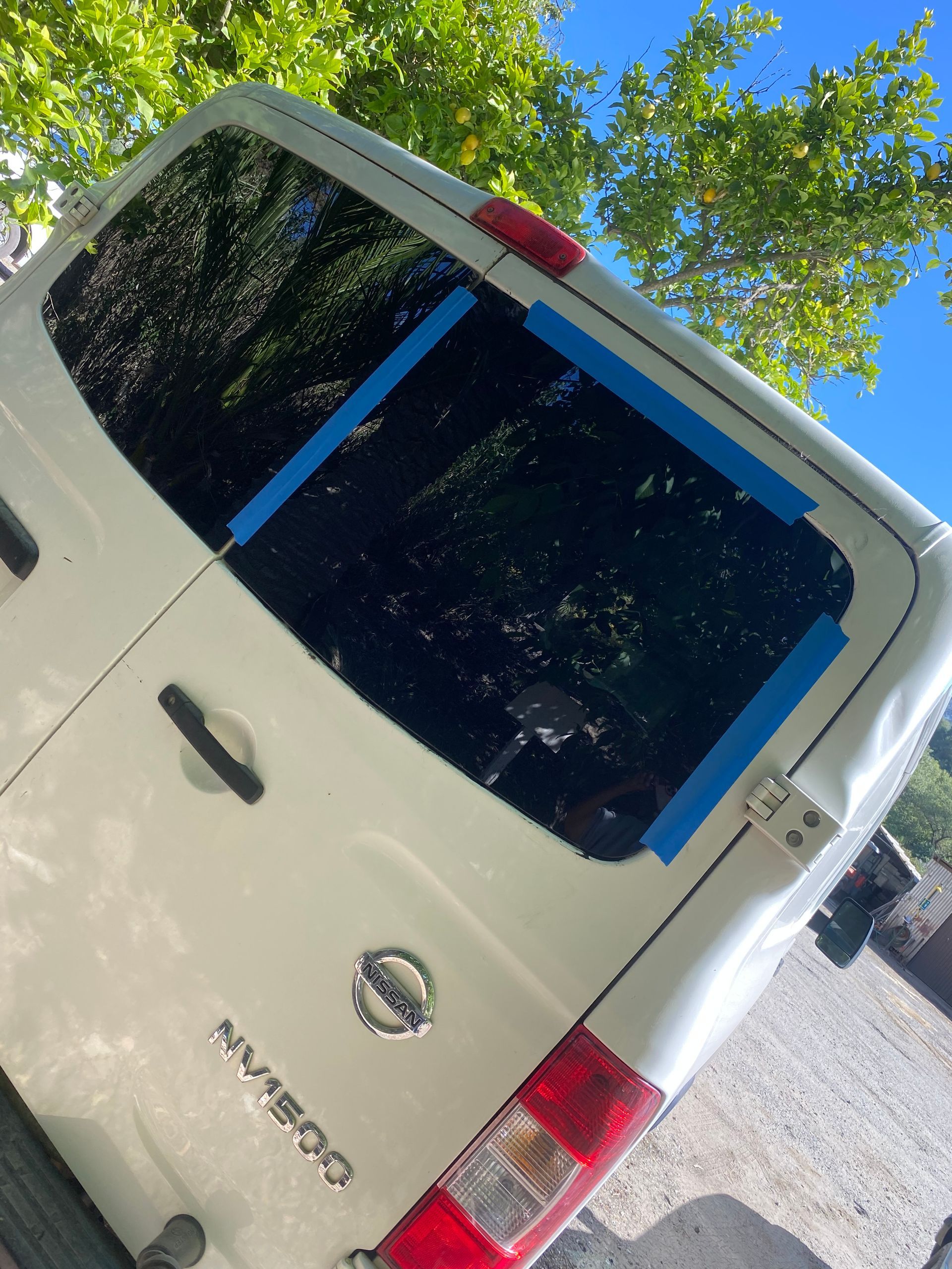 White Nissan van, back window taped, blue tape, parked outdoors with trees.