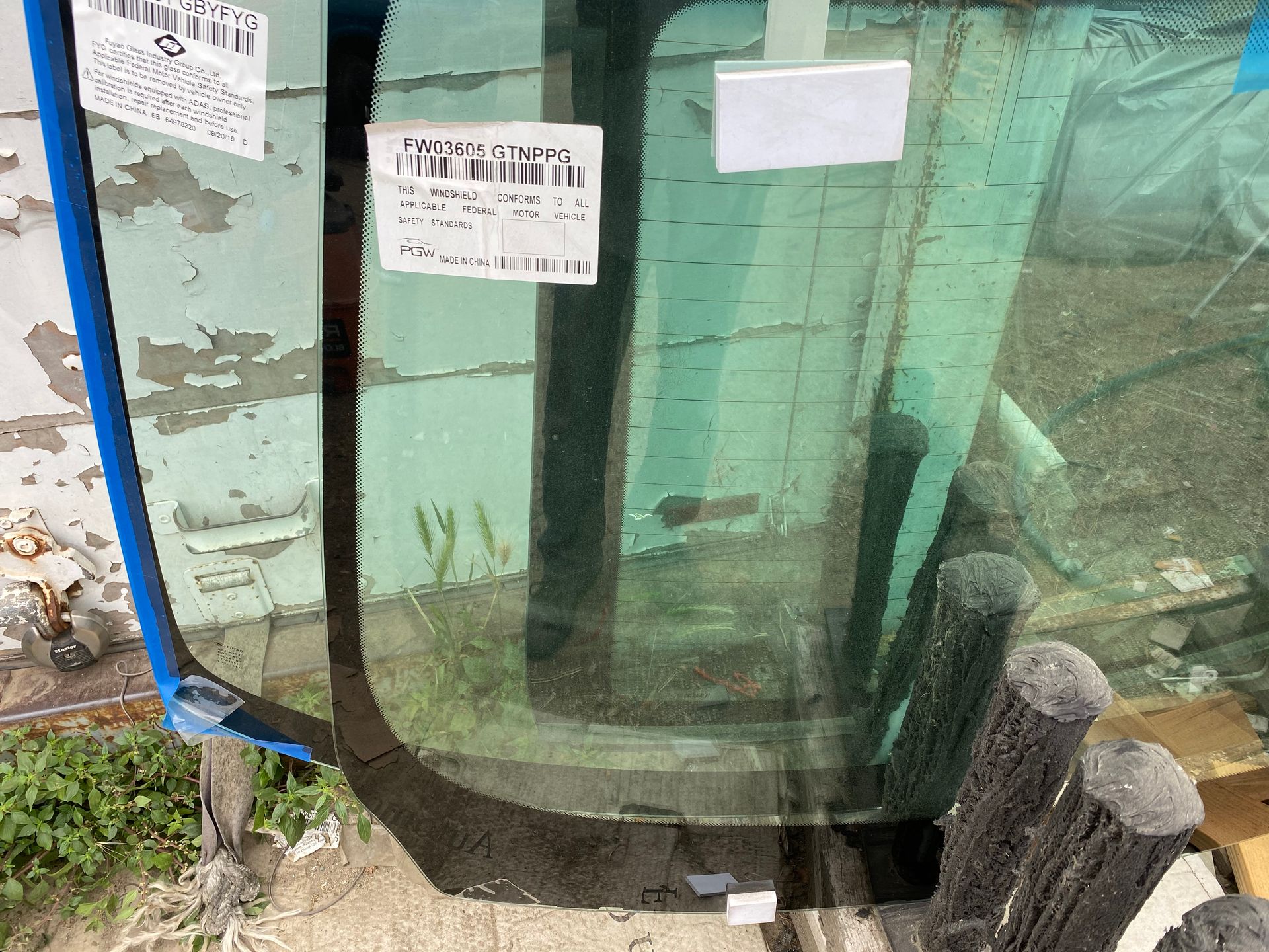 Windshield glass, with labels attached, resting against a weathered structure, outdoors.