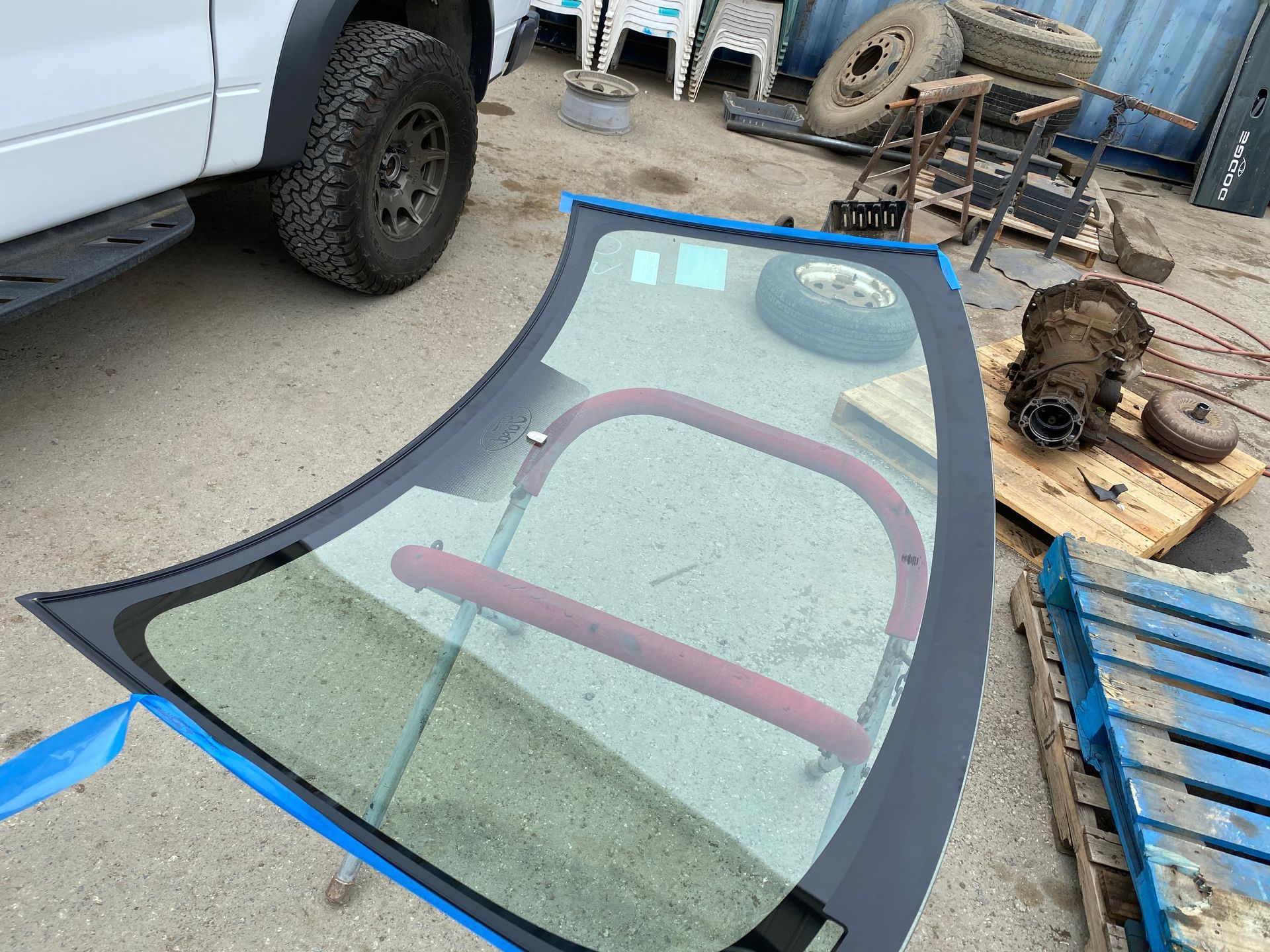 Windshield with a black frame and pink support, lying outdoors with vehicle parts in background.