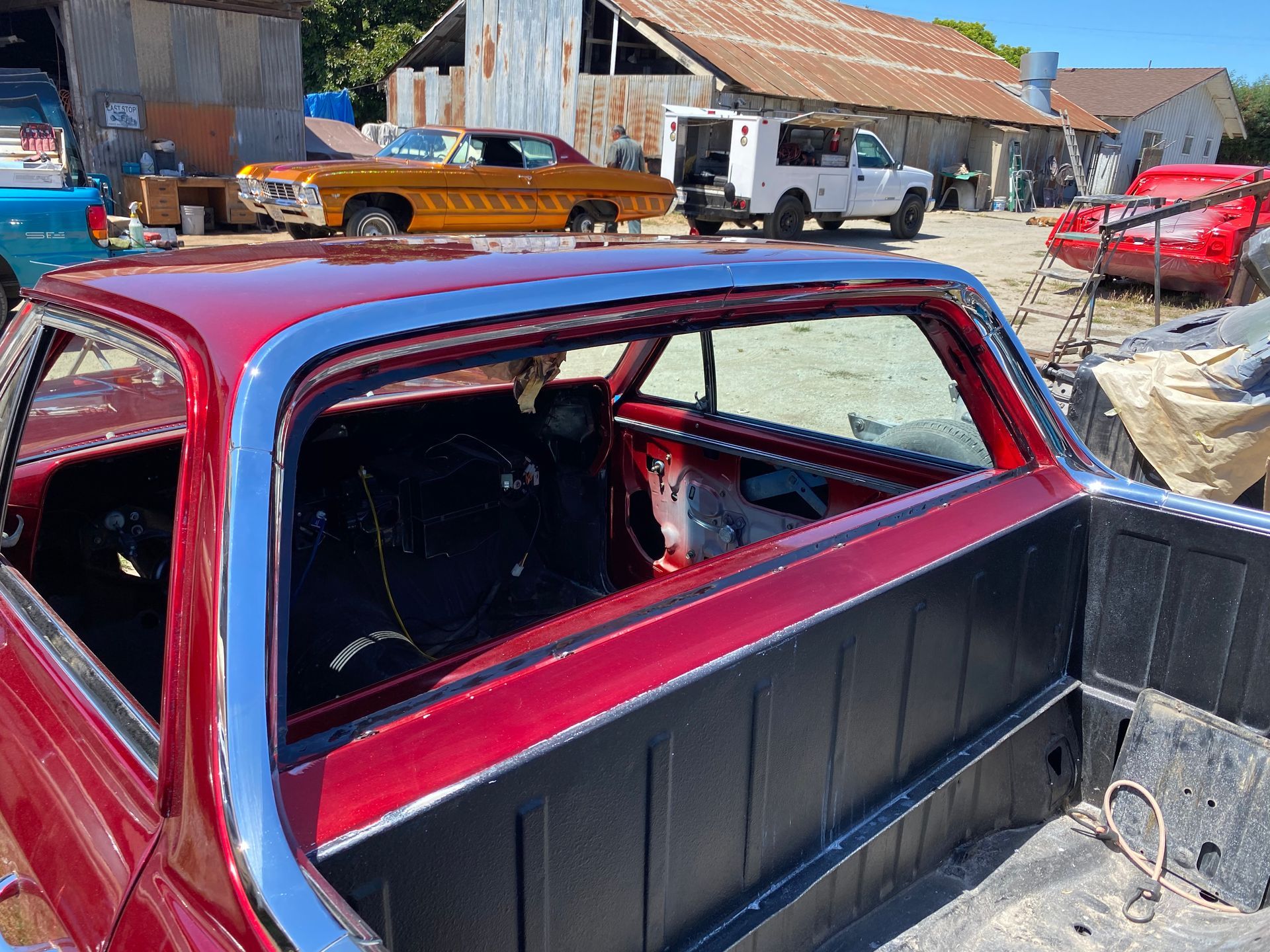 Red El Camino's interior and truck bed in a sunny workshop. Other classic cars, a barn, and a white truck are in the background.