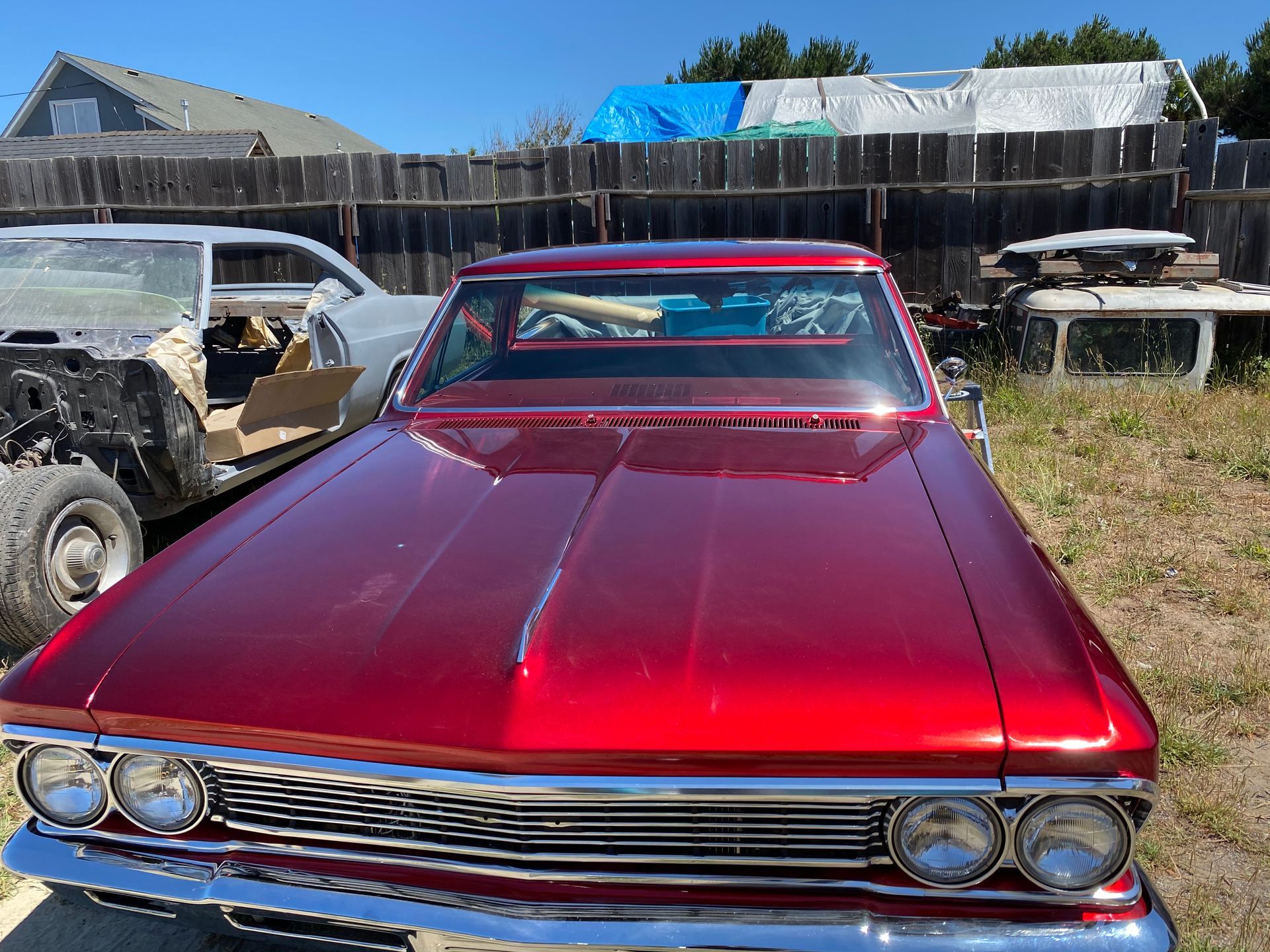 Red classic car parked in a yard with other car parts and a wooden fence.