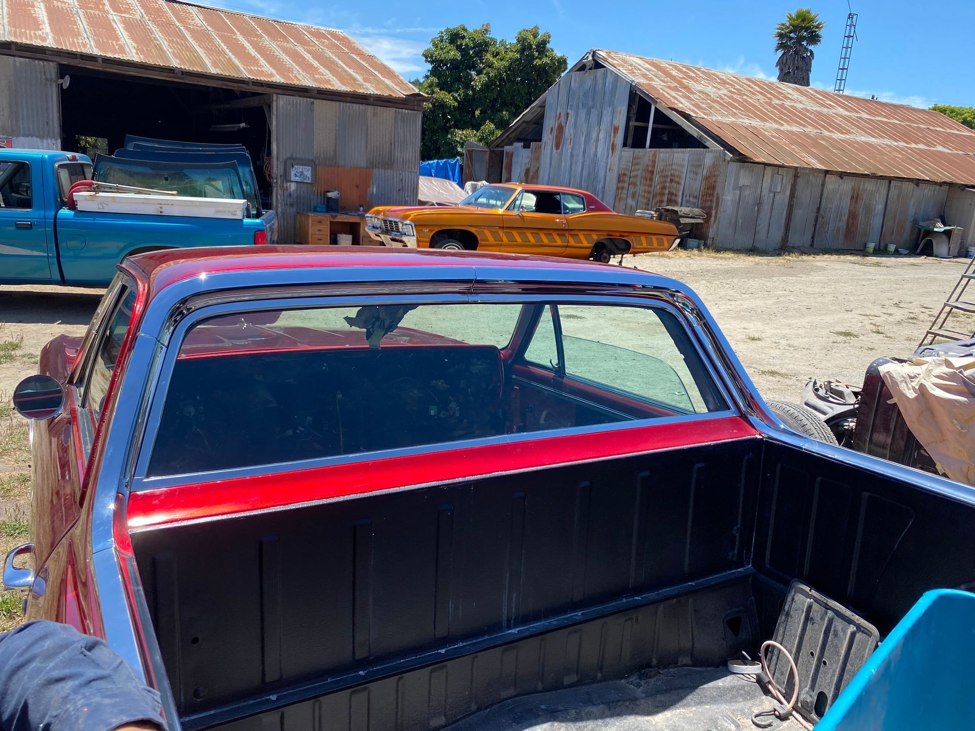 Red classic El Camino with black bed, near garages, gold car visible.