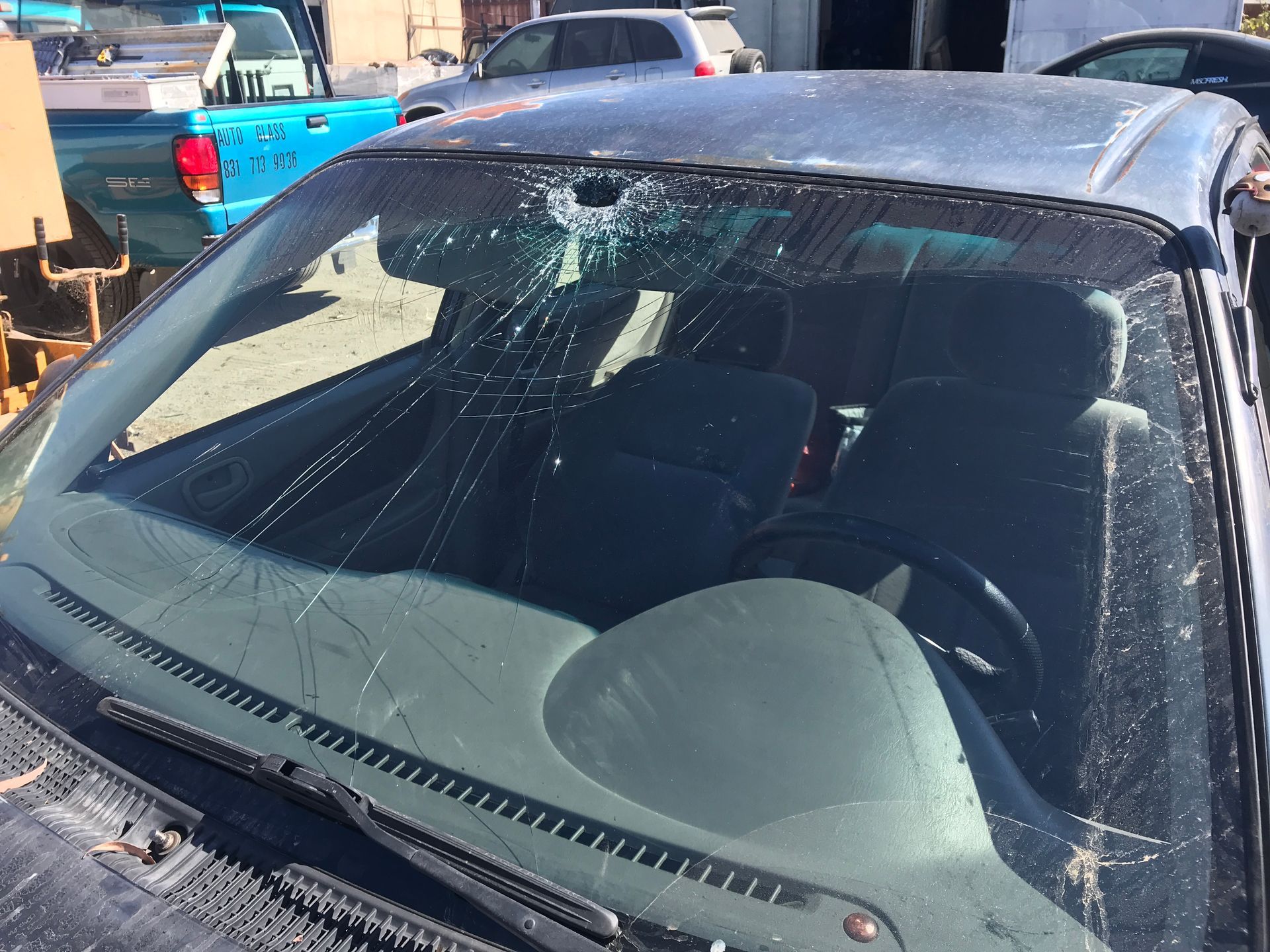 Damaged windshield of a dark-colored car, with extensive cracks. Interior seats and dashboard are visible.