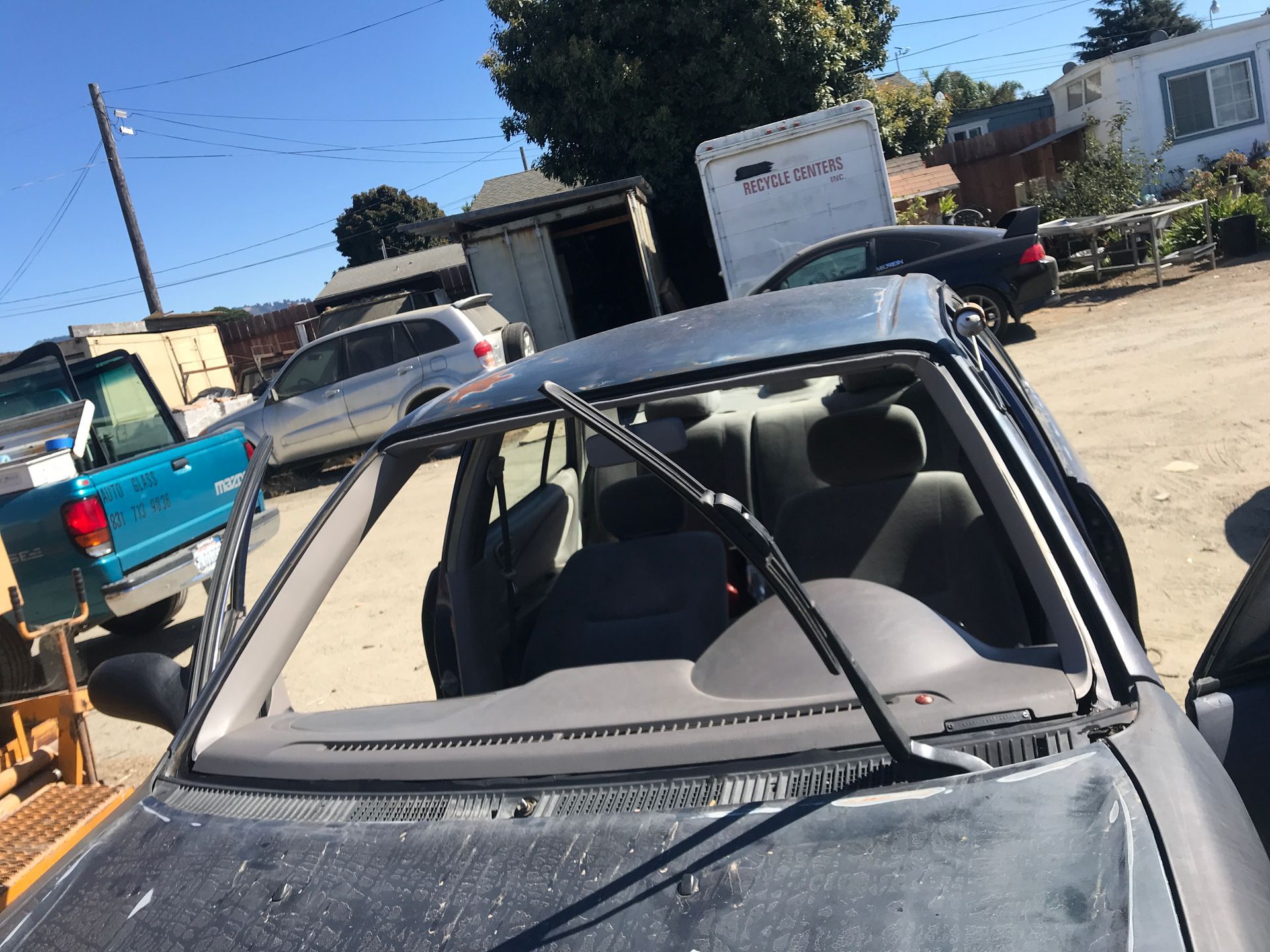 A car with a missing windshield, parked outdoors. Wiper blades are visible. Other cars and a building are in the background.