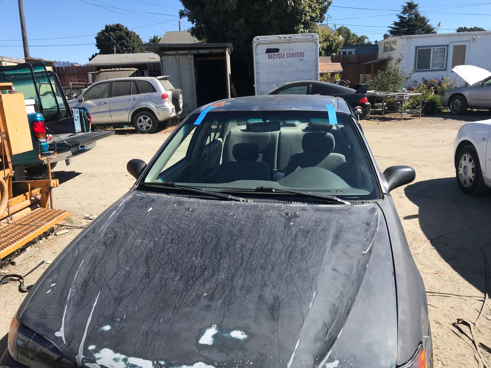 Dark car with blue tape on windshield in a sunlit outdoor lot with other vehicles and structures.