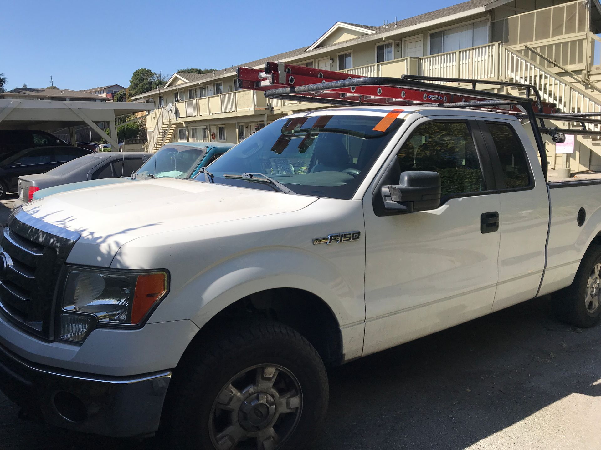 White Ford pickup truck with ladder rack parked in front of an apartment building on a sunny day.