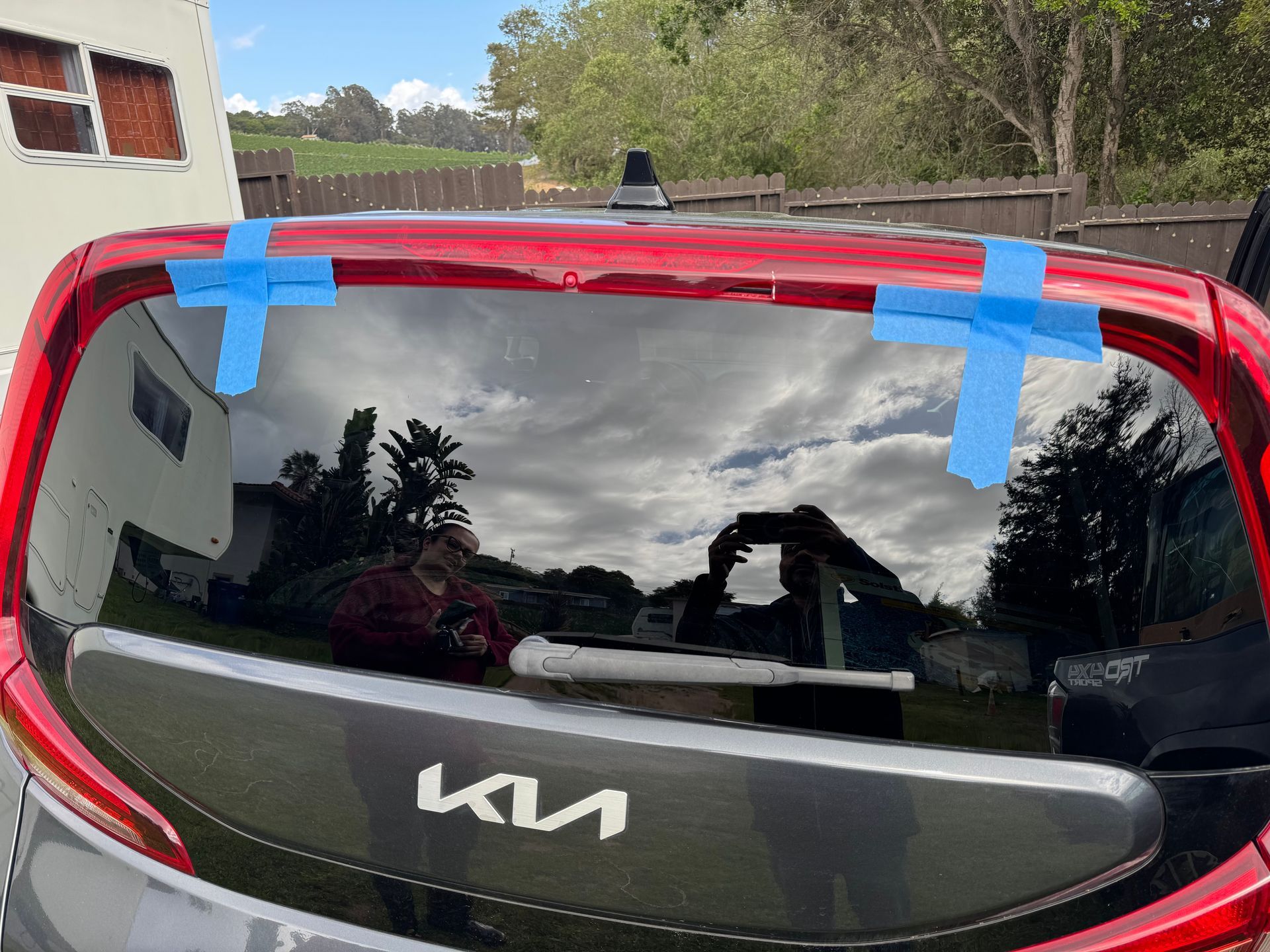 Rear of a Kia Soul with blue tape on the window, reflecting a person taking a photo and a tree-lined background.