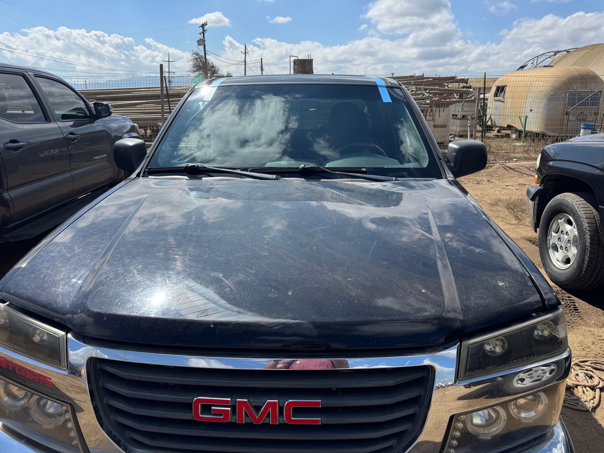Black GMC truck in a junkyard with weathered paint. The GMC emblem is red. The windshield has a blue tape.