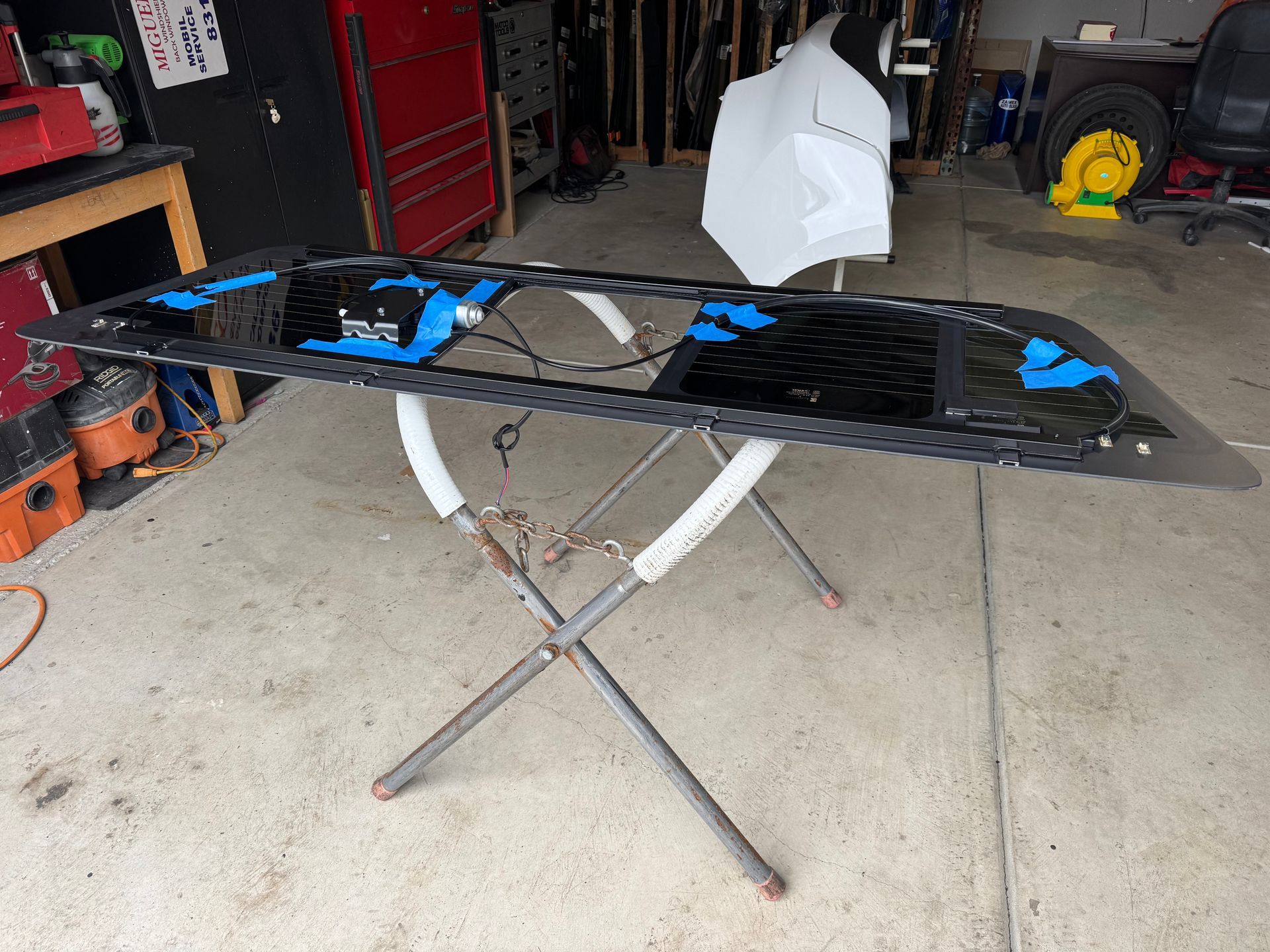 Black car part on a metal stand in a garage. Blue tape, white paint, and tools are visible.