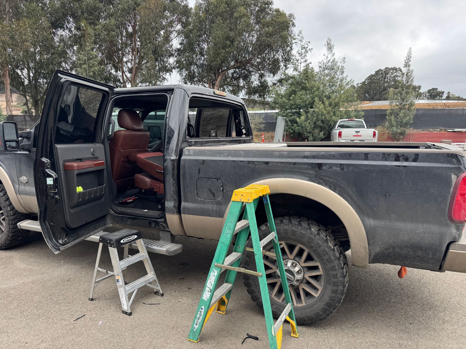 Black pickup truck with open doors, ladder, and small stool. Exterior setting.