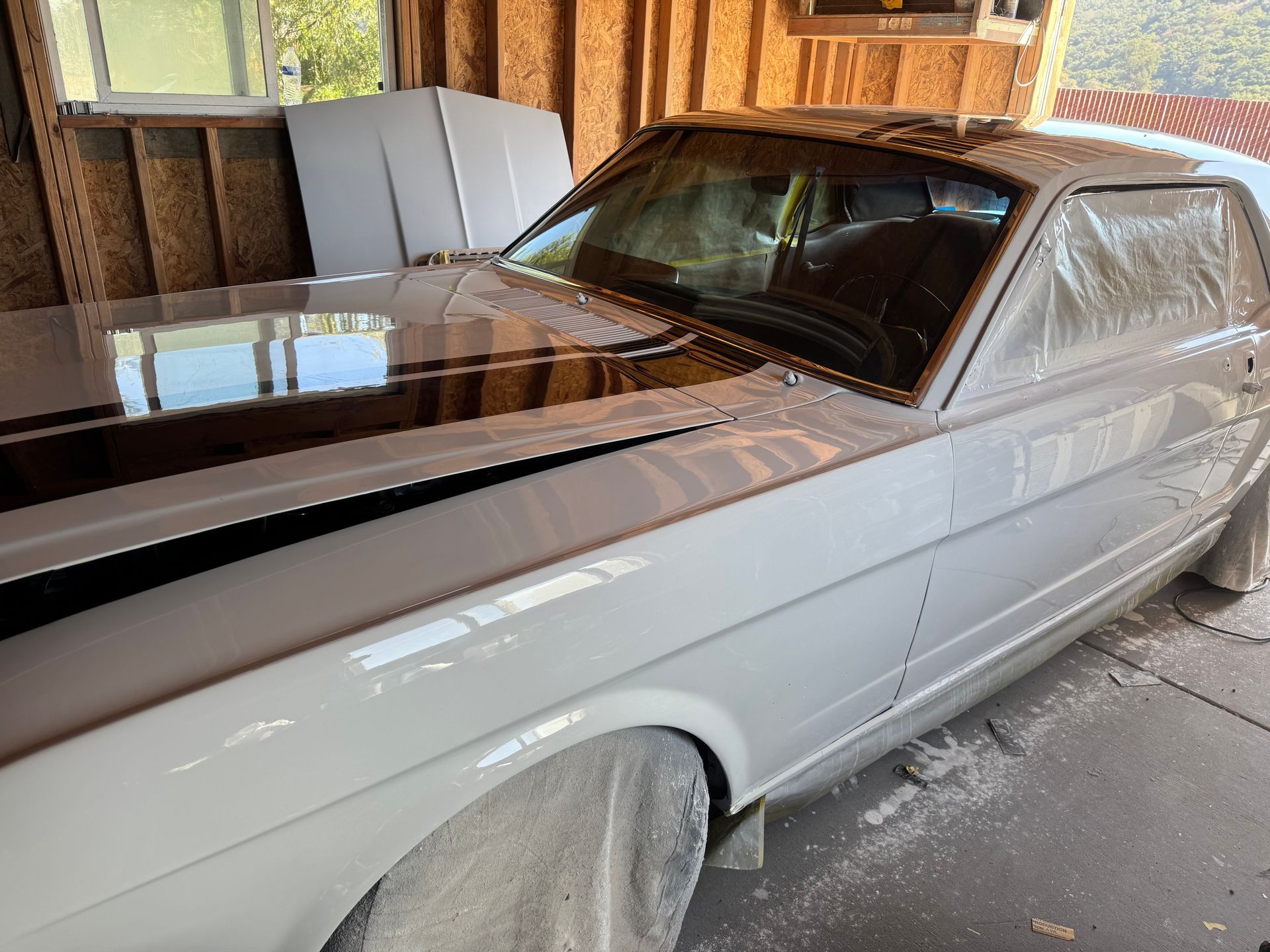 White classic car with black racing stripes on the hood, covered in plastic for painting, in a garage.
