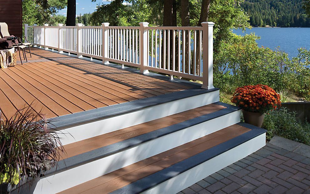 Wooden deck with white railing and steps beside a lake, with potted plants and a paved path.