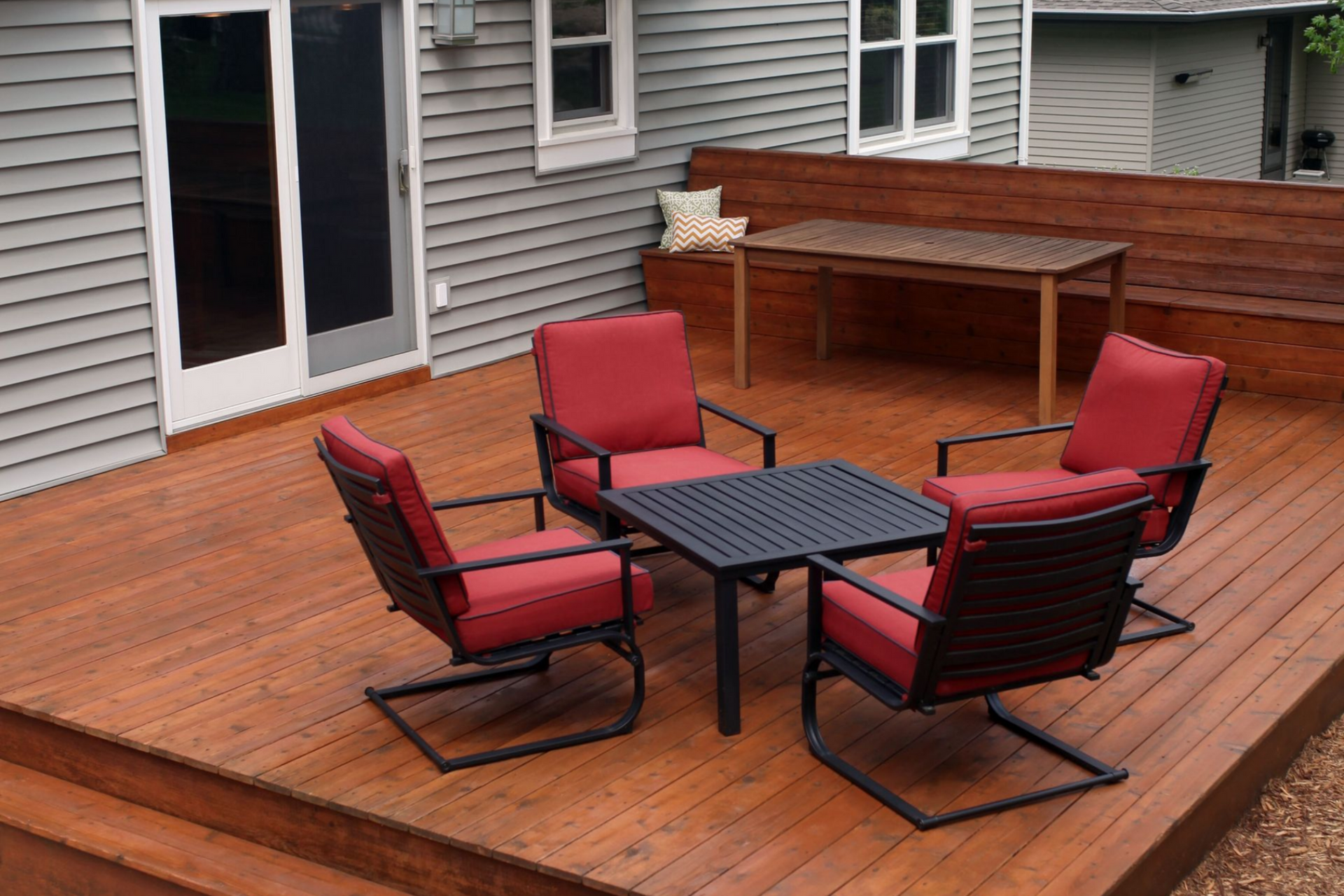 Wooden deck with four red cushioned chairs around a black table beside a house.